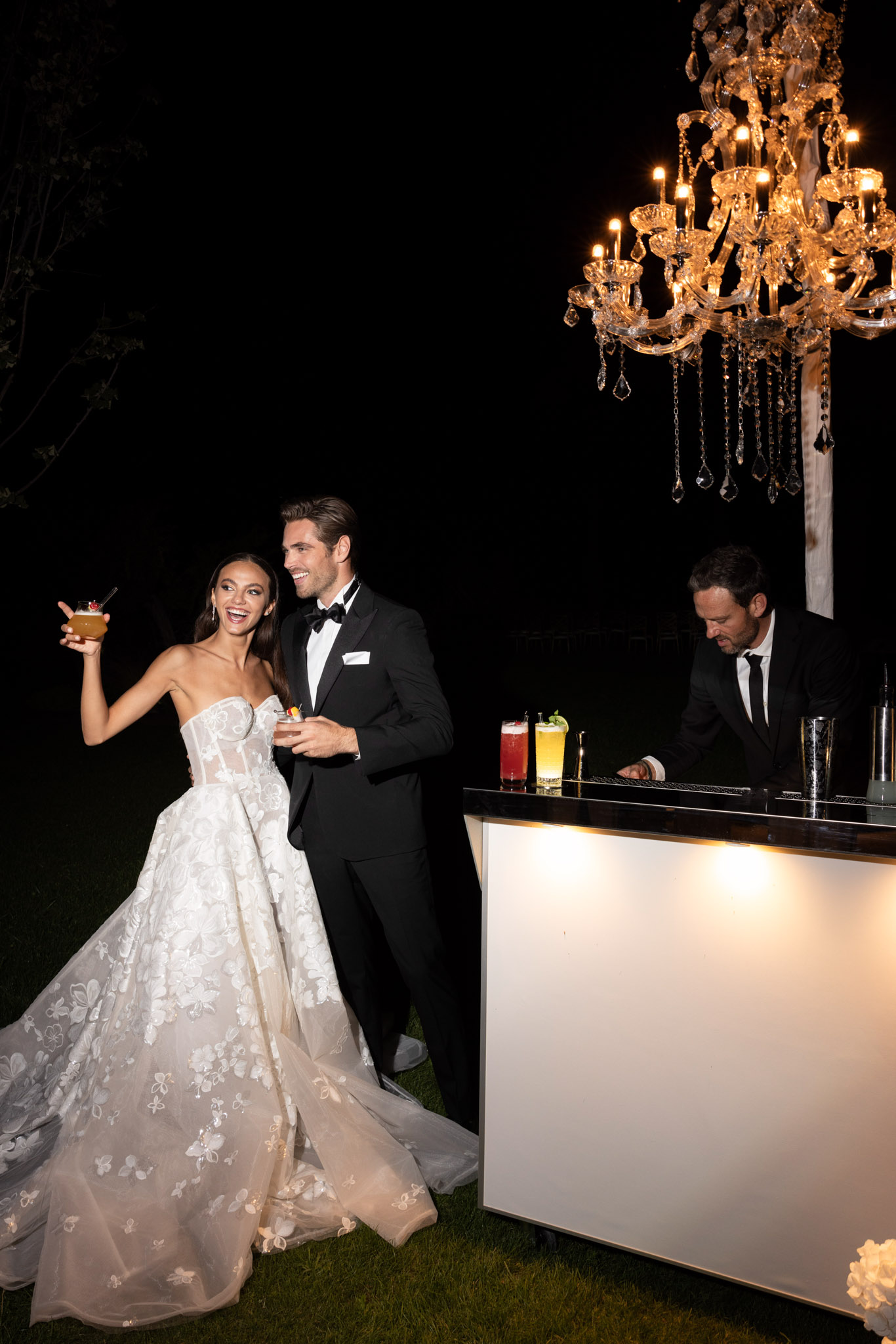 Couple holding cocktails at illuminated white bar with crystal chandelier overhead at night reception