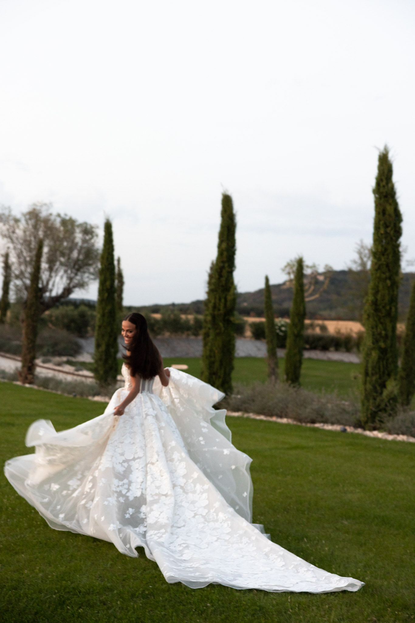 Bride twirls in floral-applique ballgown with cathedral train spread across lawn before cypress row