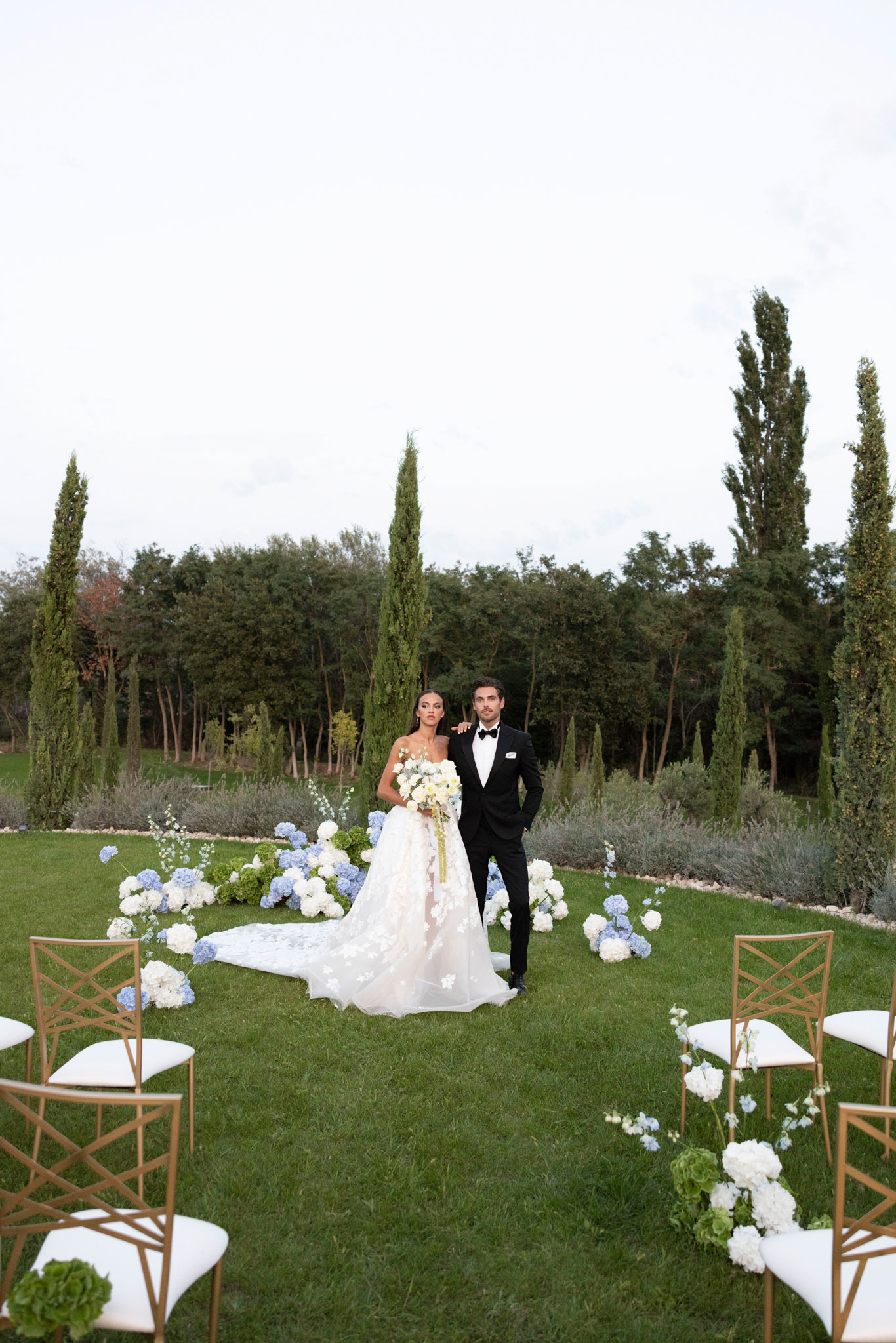 Couple at altar with crescent blue hydrangea arrangement, gold chiavari chairs, and cypress tree backdrop