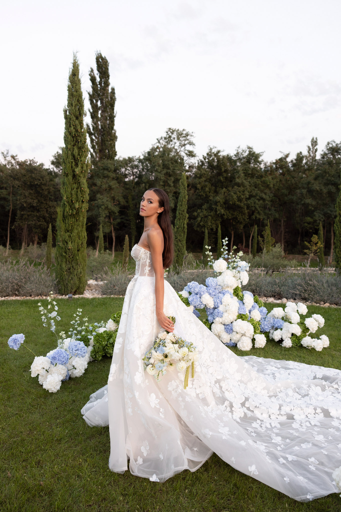 Bride in strapless floral applique ball gown with cathedral train holding ivory and blue bouquet in formal garden