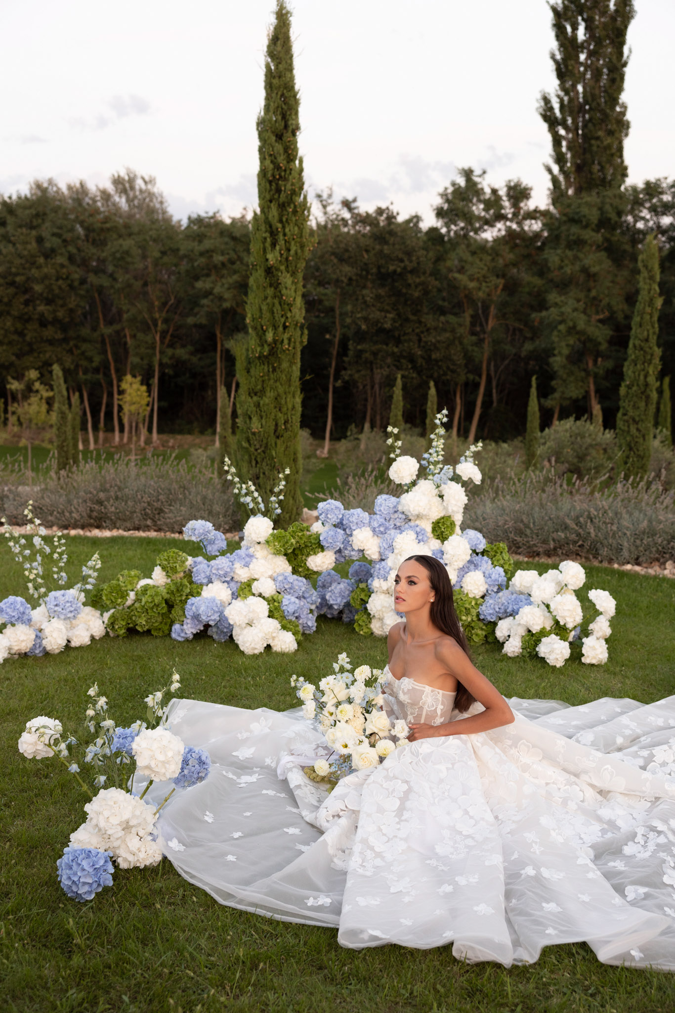 A bridal portrait taken outdoors in a formal garden setting, with the bride seated on the lawn with her full skirt and cathedral-length veil spread around her. She wears a strapless ivory ball gown with a lace-appliqué bodice and a voluminous tulle skirt embellished with three-dimensional floral or butterfly appliqués throughout. She holds a bouquet of cream/ivory roses and white delphinium. Behind her is a large ground-level floral installation composed of blue hydrangeas, white hydrangeas, and green hydrangeas arranged across the lawn, with additional stems of white delphinium rising vertically. Loose stems of blue and white hydrangeas are also placed around the spread of her skirt in the foreground. The overall color palette of the florals is blue, white, and green against the ivory gown. The composition is a medium-wide portrait shot from a slightly elevated angle, capturing the full skirt spread and the floral backdrop together.