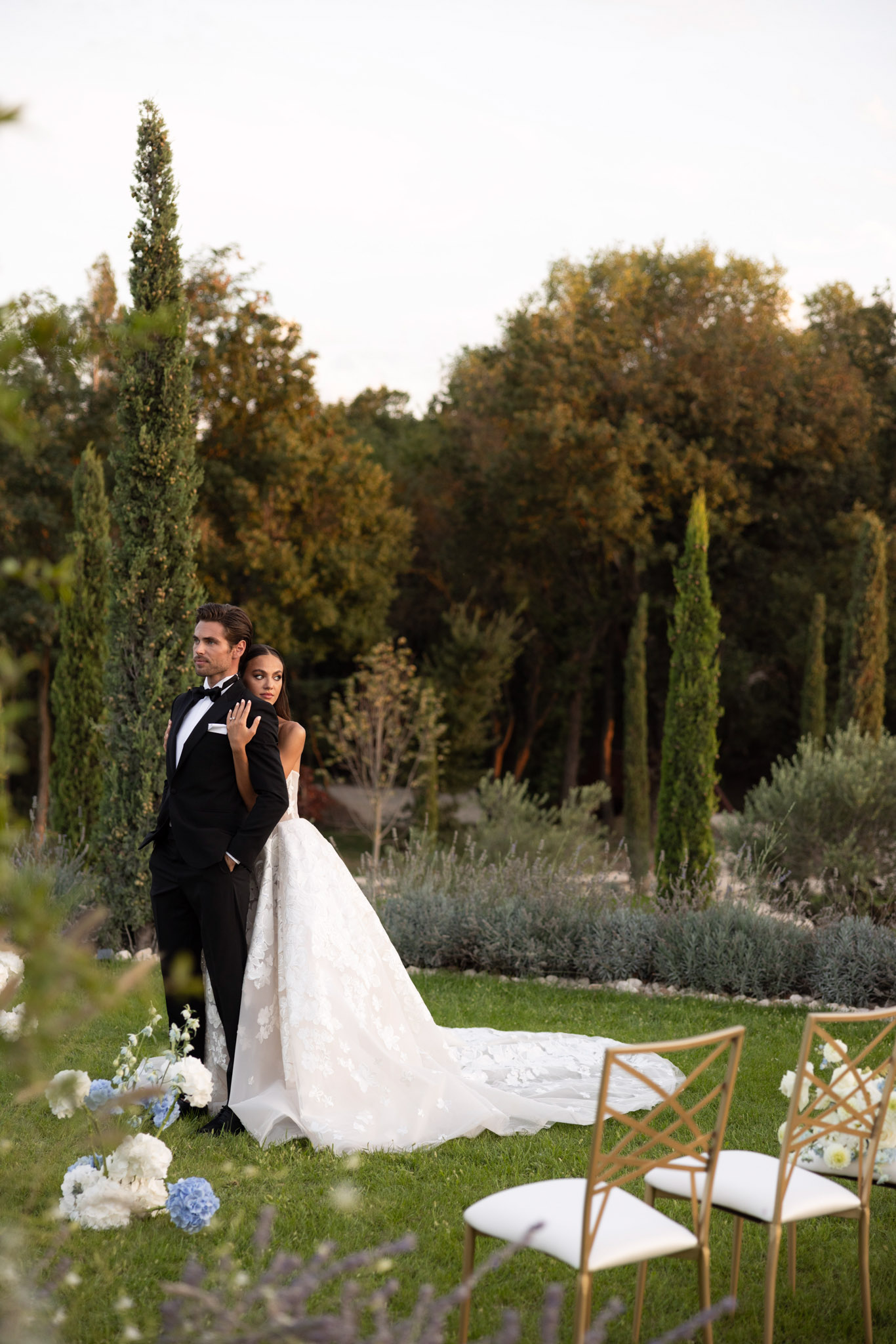 A couple portrait taken outdoors in a formal garden setting featuring tall cypress trees and manicured lavender and silver-foliage hedgerows. The bride wears a strapless ivory ballgown with floral lace appliqué and an extended cathedral train, while the groom is dressed in a black tuxedo with a black bow tie; the bride stands behind him with her hand on his chest as he looks to the side. The ceremony space is partially visible, with gold cross-back chairs with white cushions and low floral arrangements of pale blue hydrangeas and white blooms placed at ground level along the aisle. The overall styling palette is black, ivory, and powder blue, with a classic formal aesthetic, captured in a wide portrait shot at soft evening light.