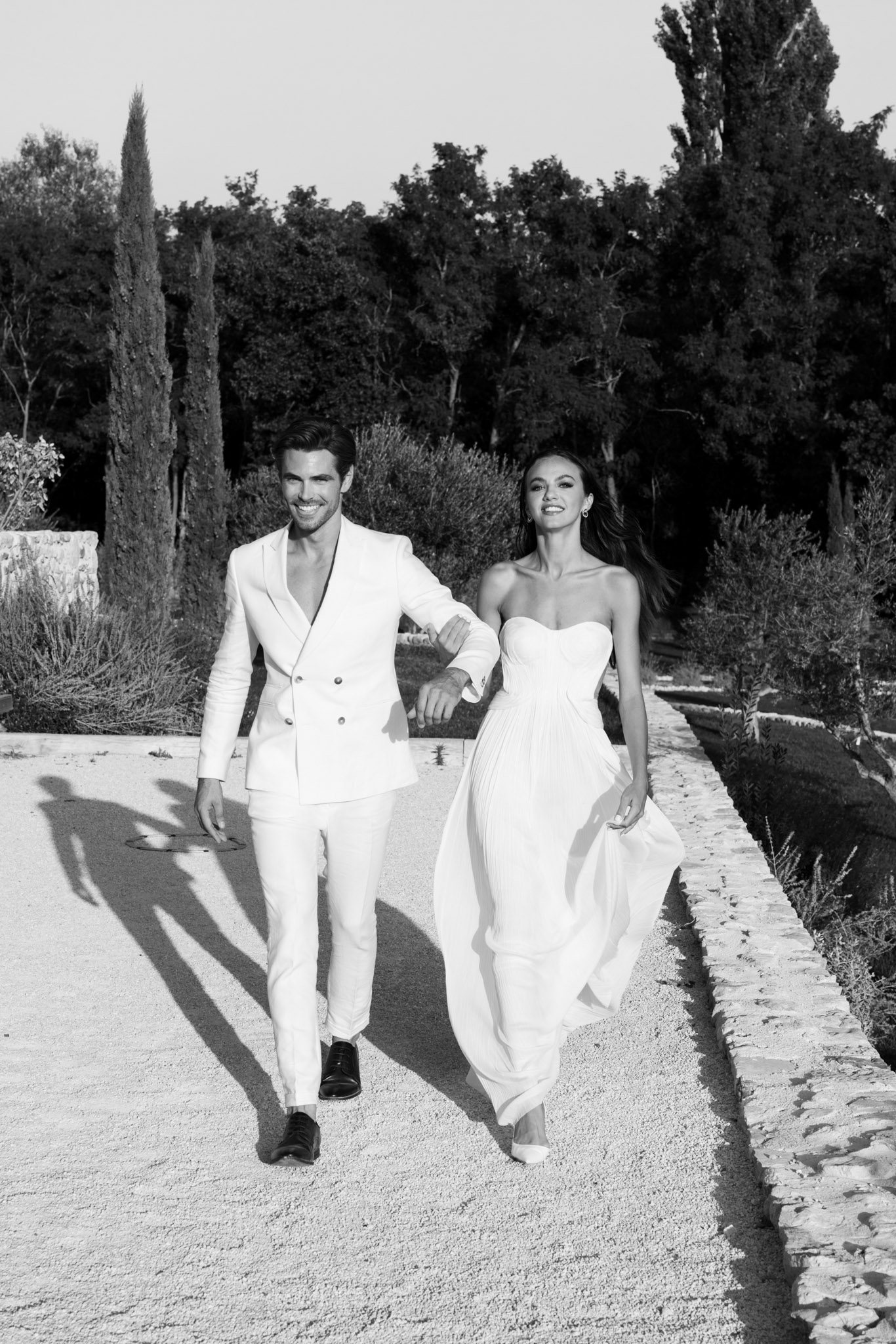 Black and white portrait of couple in all-white outfits walking arm in arm on formal garden path