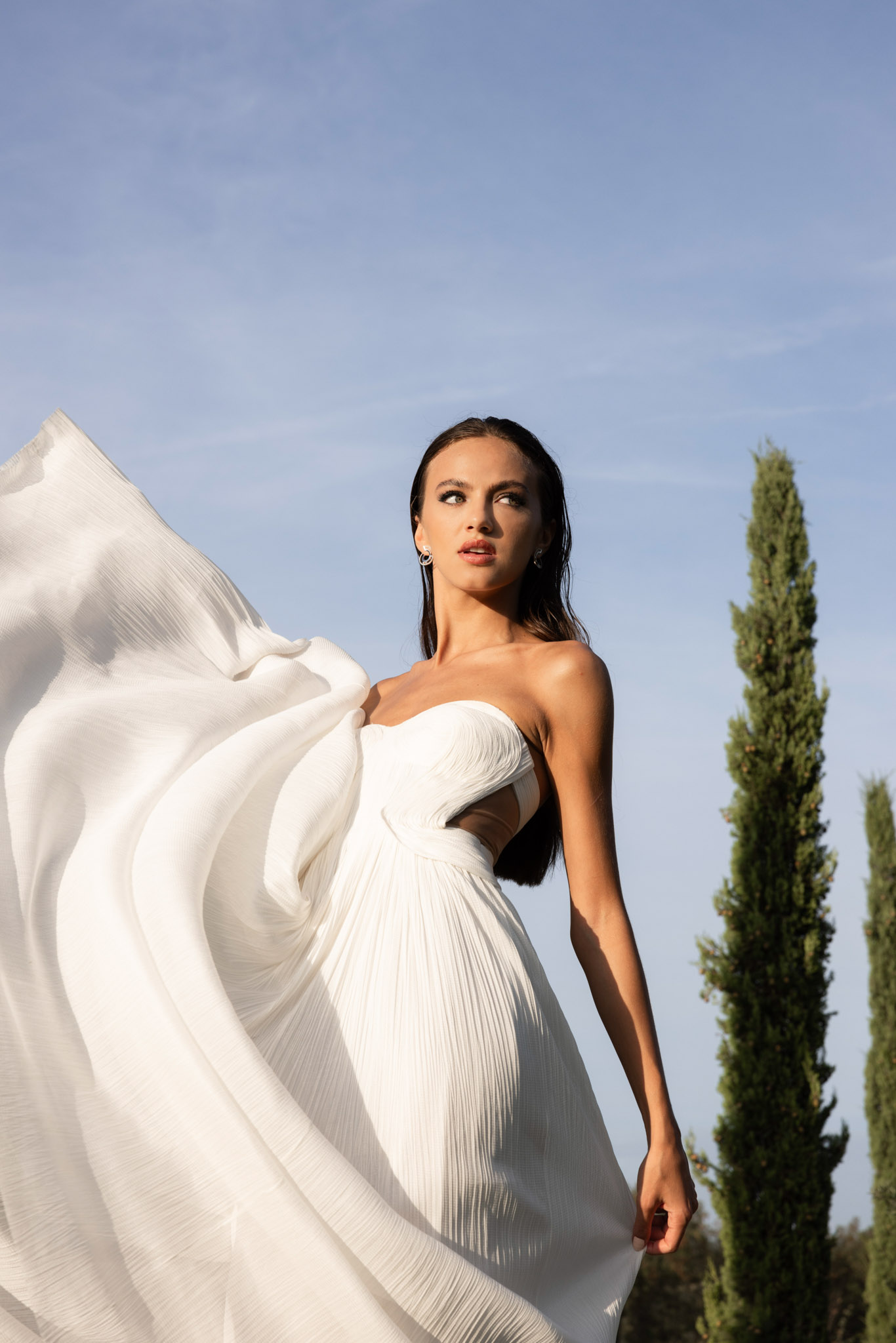 Bride in strapless pleated gown with billowing cape caught in wind, low angle against open sky with cypresses