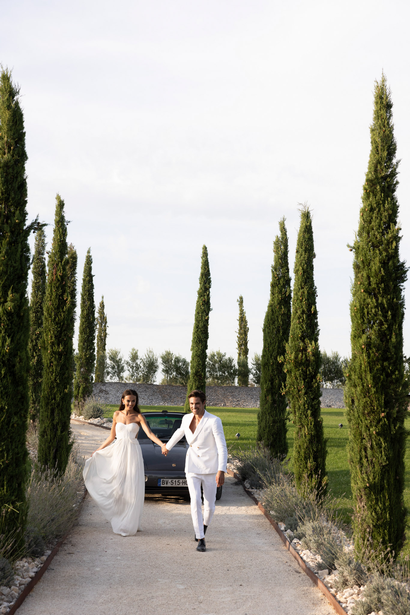 Bride and groom walking hand-in-hand along cypress-lined gravel driveway in matching white outfits