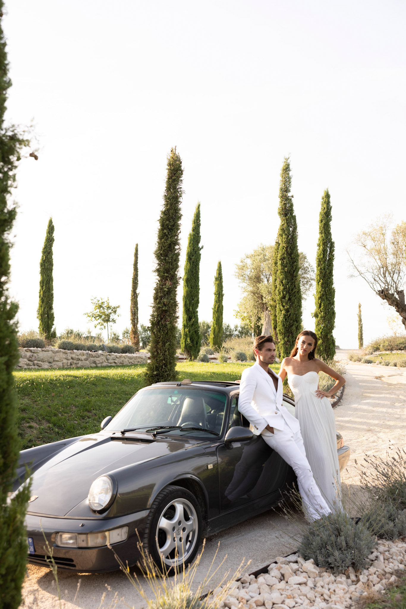 Bride and groom leaning against vintage Porsche 911 on gravel drive lined with cypress trees