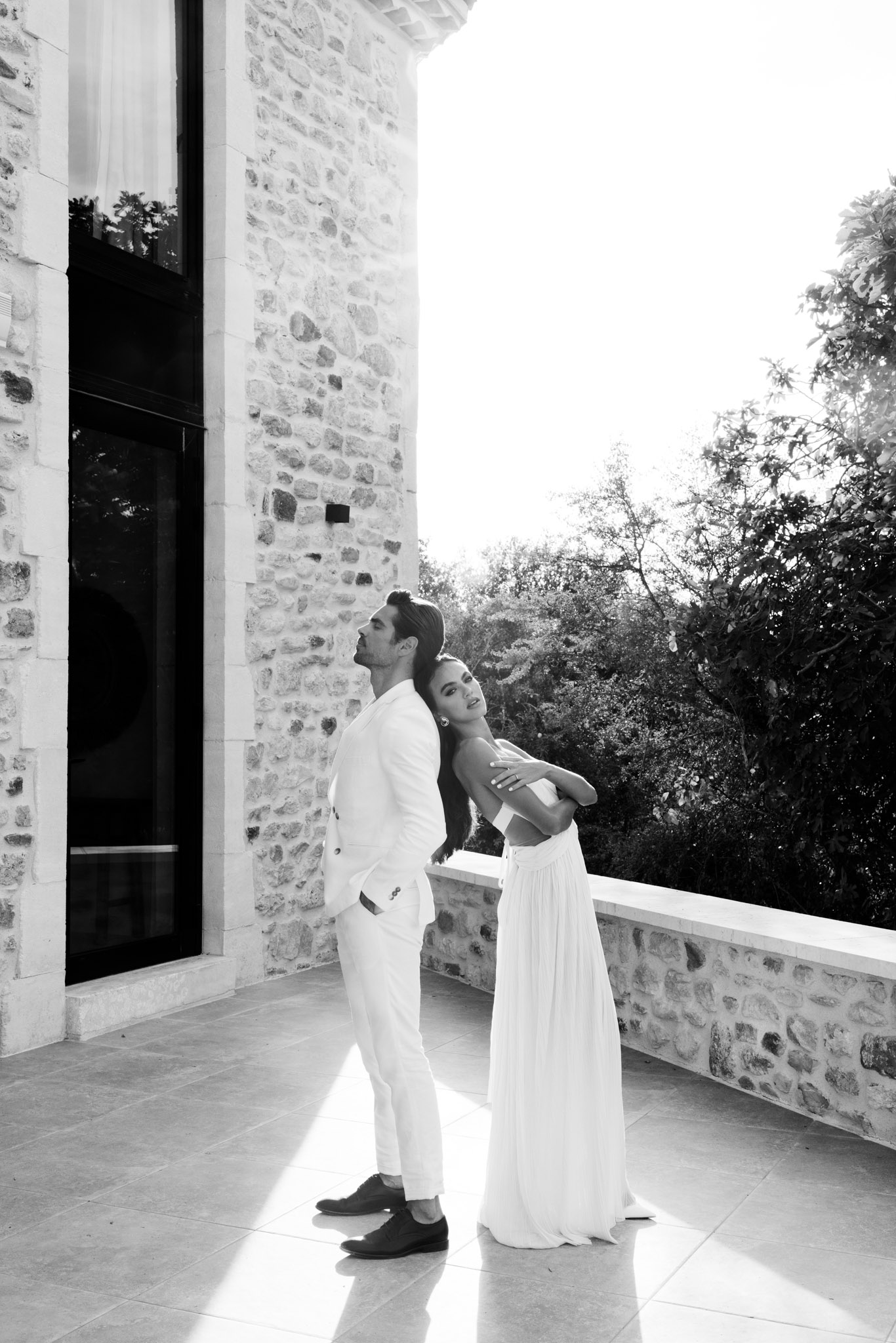 This is a black-and-white couple portrait taken outdoors on a stone terrace of what appears to be a French château or mas. The bride stands behind the groom with her arms wrapped around his torso, her head resting against his back as she looks upward; the groom faces away from her slightly, also looking upward. The groom wears a white double-breasted suit with dark Oxford shoes, and the bride wears a strapless, flowing white gown with a relaxed, draped silhouette. The image is high-contrast with bright highlights from direct sunlight casting a strong beam across the tiled terrace floor, set against the deep shadows of the stone building facade and large modern black-framed windows. The composition is a full-length portrait shot with the couple positioned to one side, the stone wall and treeline forming the background.