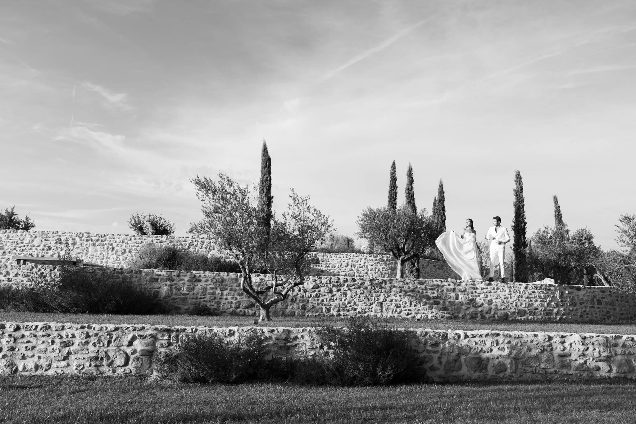 Bride with windswept cape and groom on stone terrace against terraced landscape with cypress trees in B&W