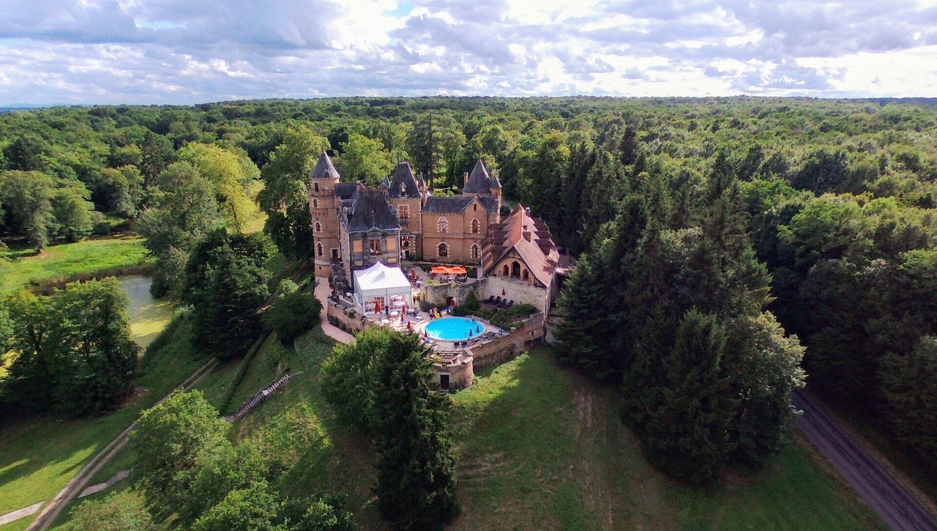 Aerial drone shot of a French château set within an extensive forested landscape, captured during what appears to be a wedding event. The château features multiple pointed turrets, warm sandstone and terracotta-tiled architecture, and a walled courtyard. A large white reception marquee tent has been erected in the courtyard beside the château, accompanied by orange parasols and a round above-ground pool with bright blue water. A small number of guests are visible around the pool and courtyard area. A pond or small lake is visible to the left of the property, and a road borders the right edge of the grounds. Potential venue feature image.