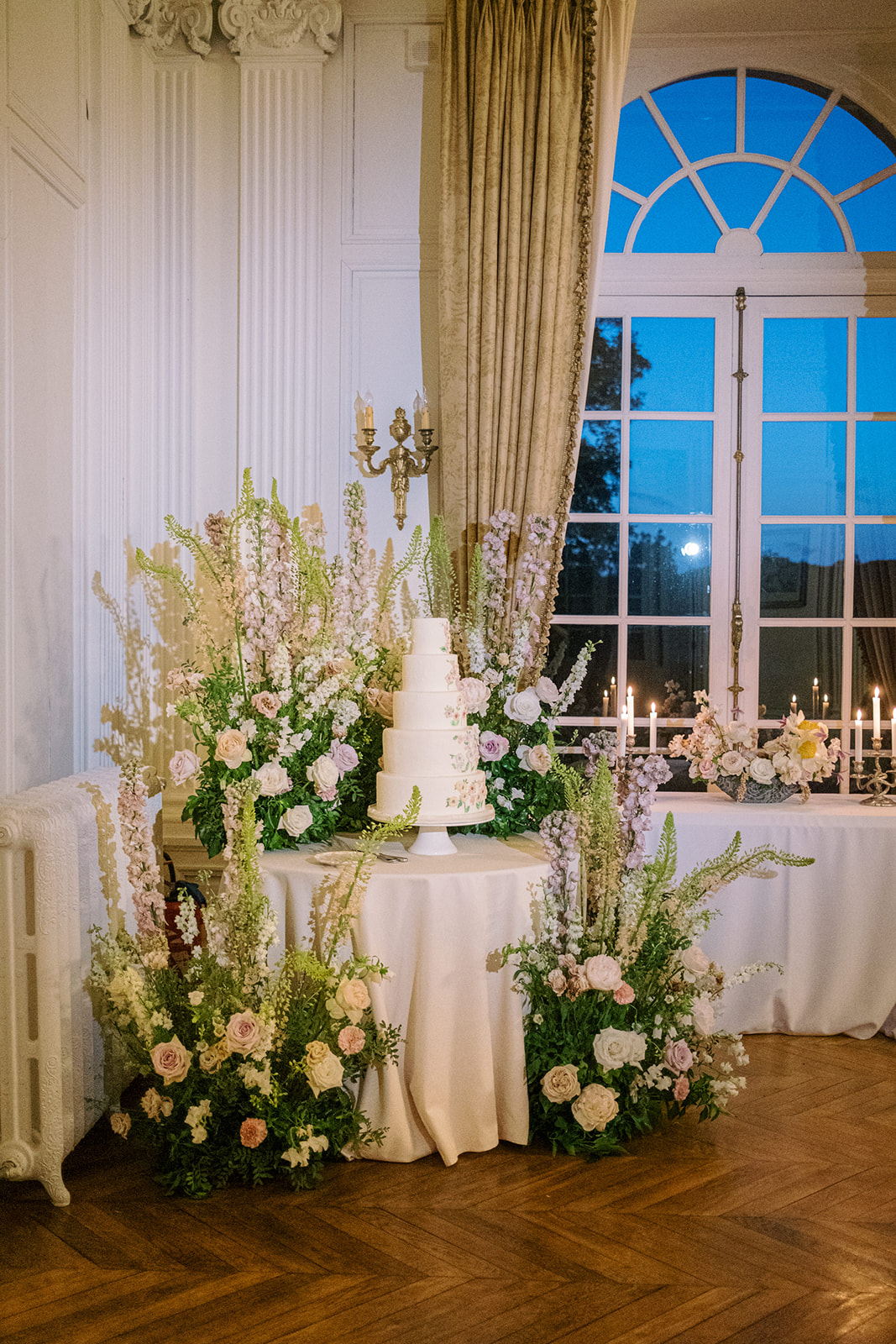 A detail shot of a wedding cake display set up inside a formal reception room, photographed in the evening with deep blue sky visible through a large arched window. The four-tier white cake sits on a white cake stand atop a round table draped in an ivory linen, and features delicate hand-painted floral motifs in soft blush and green tones. The cake is surrounded by an elaborate floral installation composed of blush and mauve garden roses, lavender and white delphiniums, pale peach ranunculus, and cascading greenery including ferns and foliage, arranged both on the table and in floor-level clusters that extend outward onto the herringbone parquet floor. In the background, a silver candelabra with lit taper candles and a small floral arrangement in similar tones are visible on a separate white-draped table. The room features white boiserie panelling, a gilt wall sconce, and gold damask curtains, consistent with a classic French château or manor house ballroom. The overall floral palette — blush, mauve, ivory, and soft green — and the garden-style, unstructured floral design give the display a romantic, botanical aesthetic.
