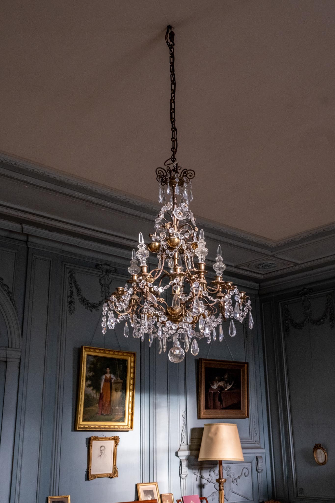 Brass and crystal chandelier in chateau room with blue-grey paneling, carved mouldings, and gilt-framed paintings