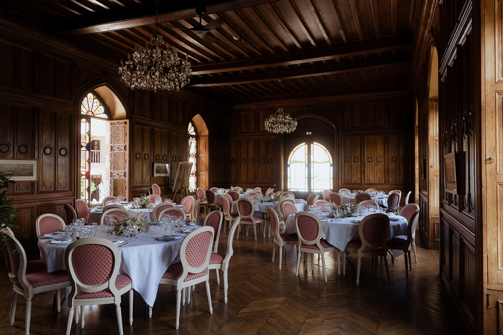 Indoor chateau reception room with dark wood paneling, crystal chandeliers, round tables, and dusty rose medallion chairs