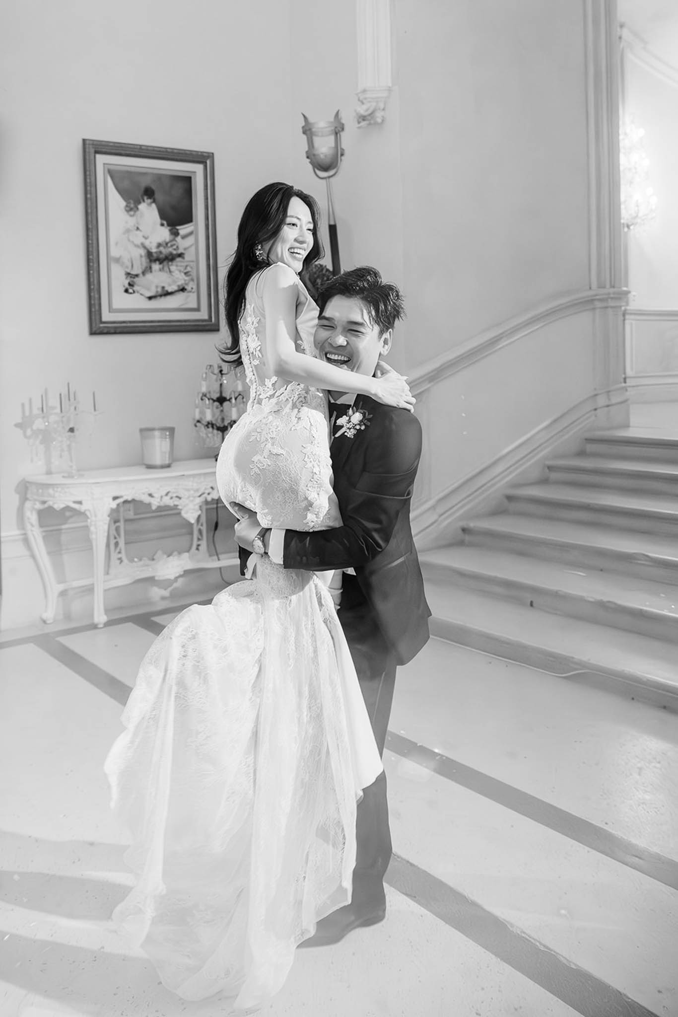 Black and white groom lifting laughing bride in lace mermaid gown in chateau hall with curved staircase and wainscoting