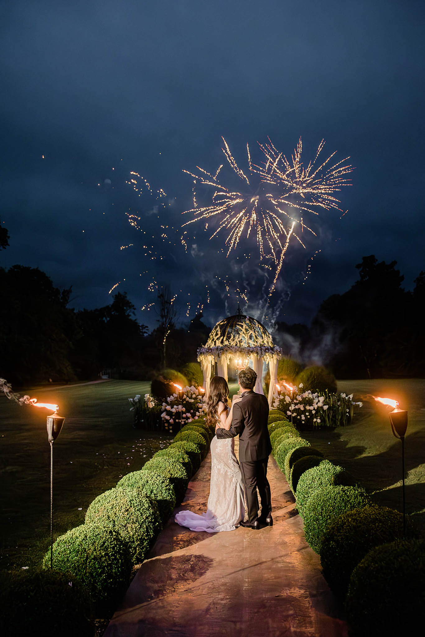 Couple watches fireworks from a torch-lit stone pathway leading to a floral-draped gazebo in a formal garden at night