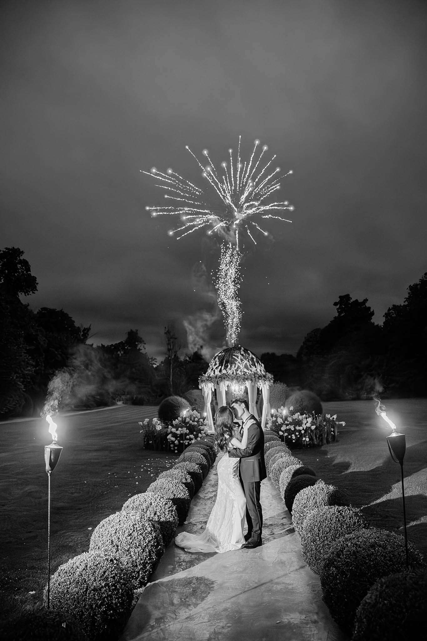 This black-and-white nighttime portrait shows the bride and groom sharing a kiss outdoors in a formal garden setting, framed by a symmetrical pathway bordered by low clipped hedging and lit tiki torches on either side. Behind them stands a decorated gazebo or rotunda adorned with florals and draped fabric, from which a firework is launching and bursting dramatically in the sky above. The bride wears a fitted gown with a train, and the groom is in a dark suit. The high contrast of the B&W image emphasizes the bright burst of the firework against the dark cloudy sky, the flickering torch flames, and the soft lighting of the gazebo structure. The wide-shot composition places the couple at the center of the garden's central axis, with the firework display serving as a deliberate visual focal point directly above them.