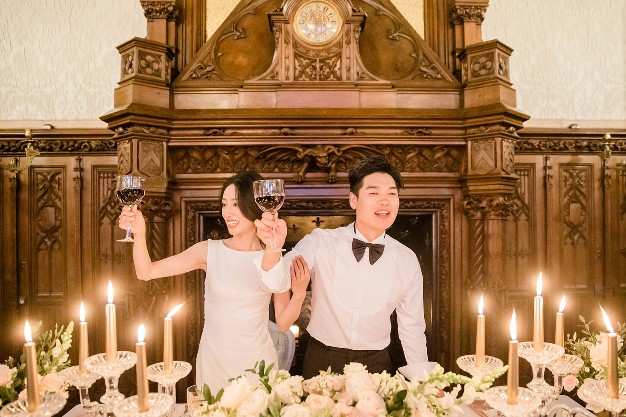 Couple toasting red wine at sweetheart table with crystal candelabras before carved Gothic fireplace