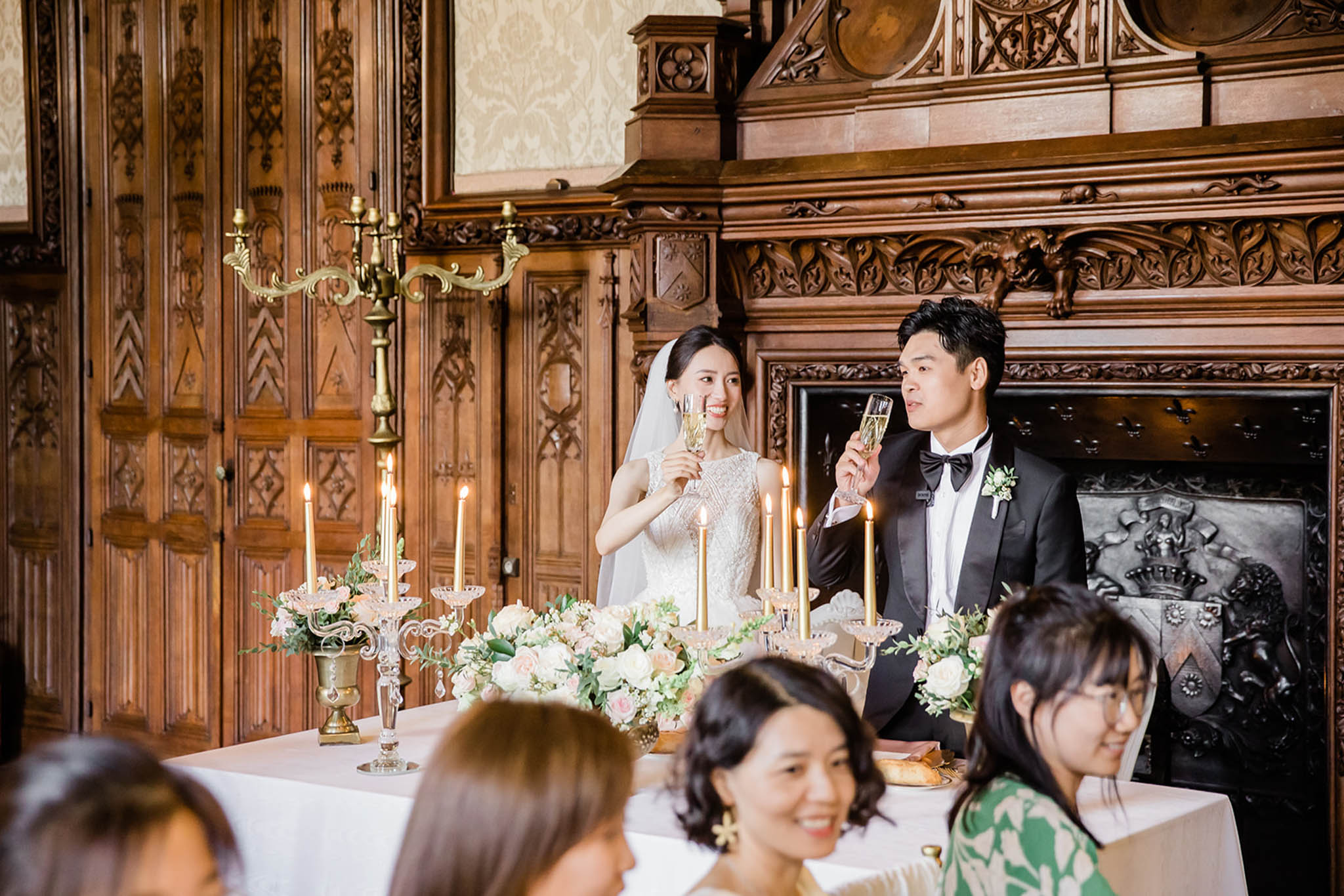 The bride and groom are raising champagne glasses during a toast at their indoor reception, seated at a sweetheart table in front of an ornate dark wood fireplace with elaborate carved detailing. The bride wears a sleeveless lace gown with a cathedral veil, and the groom is dressed in a black tuxedo with a black bow tie and a white floral boutonniere. The sweetheart table is decorated with gold candelabras holding lit taper candles and low floral arrangements featuring ivory and blush pink roses with greenery. Several guests are visible in the foreground, slightly out of focus, watching the toast. The setting is a château or manor ballroom with richly carved wood-paneled walls and damask wallpaper, styled in a classic, formal aesthetic. Medium-distance portrait-style shot.