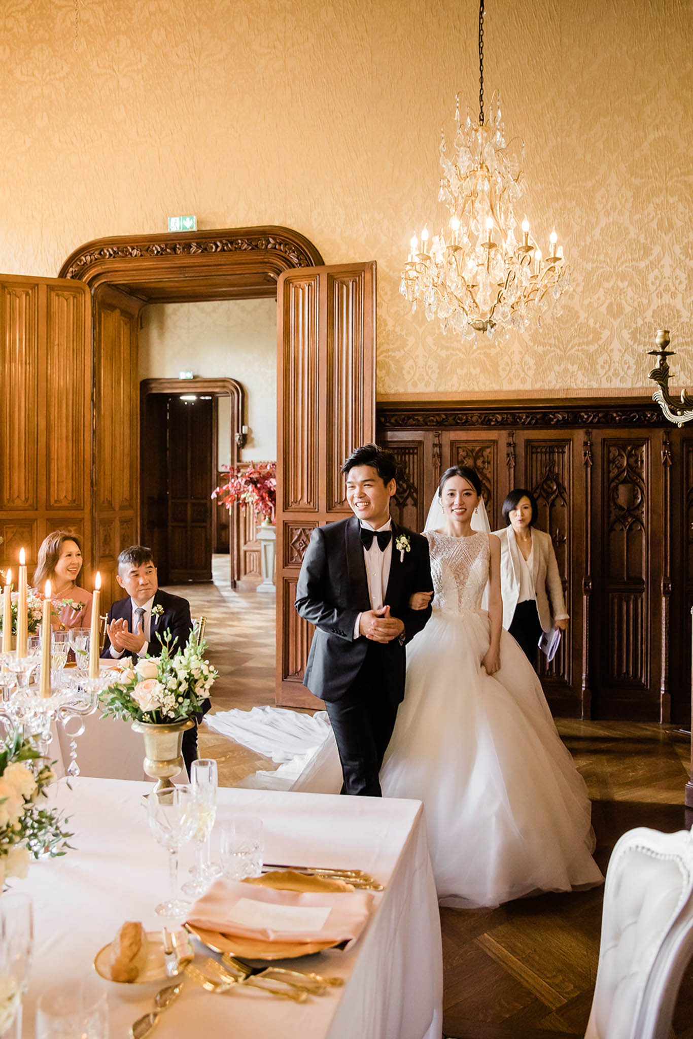 The bride and groom make their grand entrance into a formal indoor reception room, walking together through an ornate carved dark wood doorway as seated guests applaud. The groom wears a black tuxedo with bow tie and a cream boutonniere, while the bride wears a ballgown with a beaded illusion bodice, full tulle skirt, and a veil. A woman in a cream blazer follows closely behind them. The reception room features gold damask wallpaper, dark wood paneling, a crystal chandelier overhead, and herringbone parquet flooring, with a classic and formal decor style. In the foreground, reception tables are set with white linens, blush peach napkins, gold cutlery, crystal glassware, silver candelabras, and low floral centerpieces of pale peach roses and greenery; tall candles are lit along the table. Wide-angle shot capturing both the couple's entrance and the table setting in the foreground.