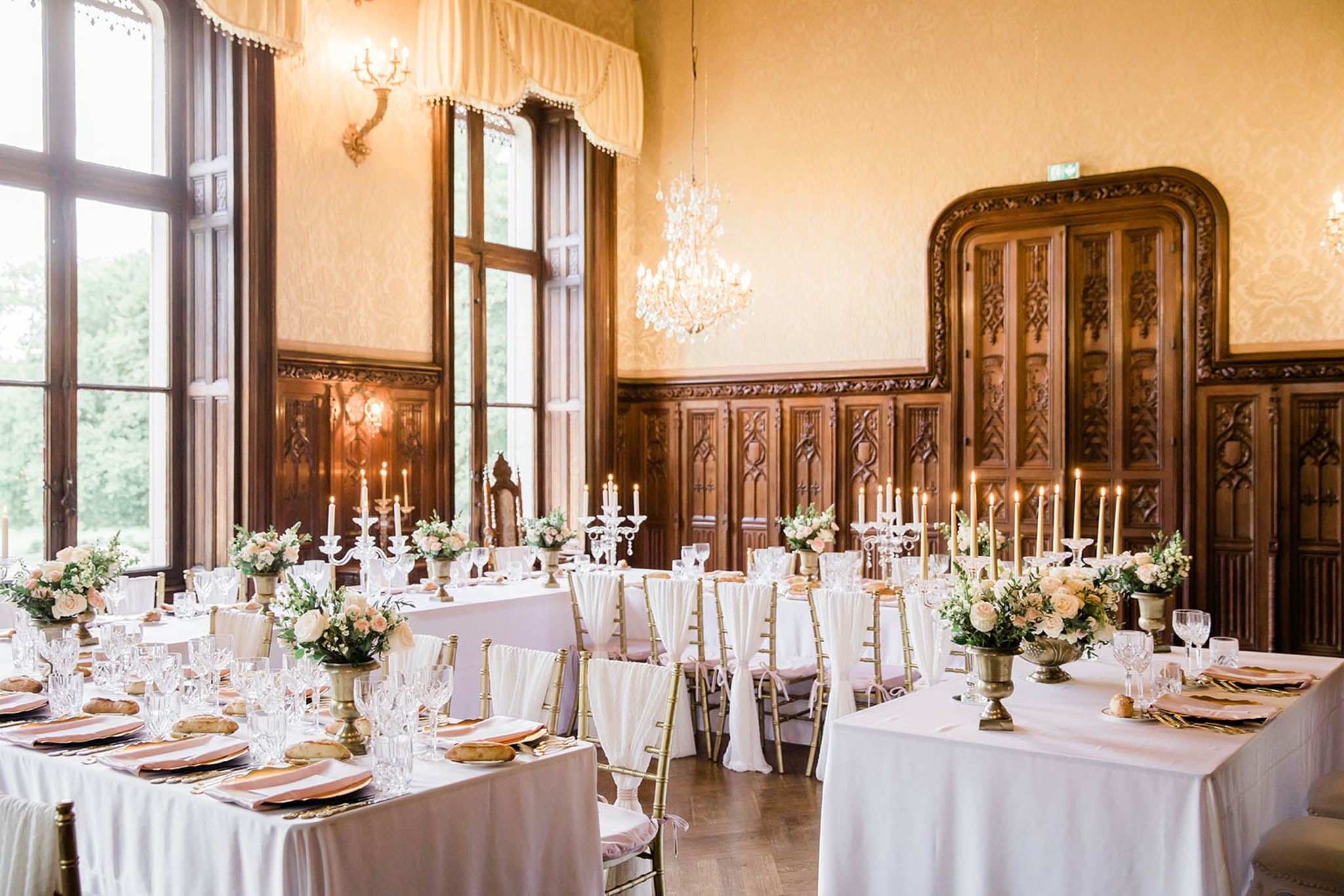 A wedding reception room set up inside a château ballroom featuring ornate dark walnut carved wood paneling, a crystal chandelier, and tall arched windows with draped cream valances. Multiple long rectangular tables are dressed in white floor-length linens with gold chiavari chairs fitted with blush pink cushions and white sash ties. Table settings include gold charger plates, blush pink linen napkins, crystal glassware, and gold cutlery. Centerpieces consist of blush and cream roses with white blooms and greenery arranged in antique gold urn vases, paired with tall white crystal candelabras holding lit ivory taper candles. The overall decor palette is white, blush pink, and gold with a classic formal style. Wide-angle interior shot showing the full room layout. Potential venue feature image.