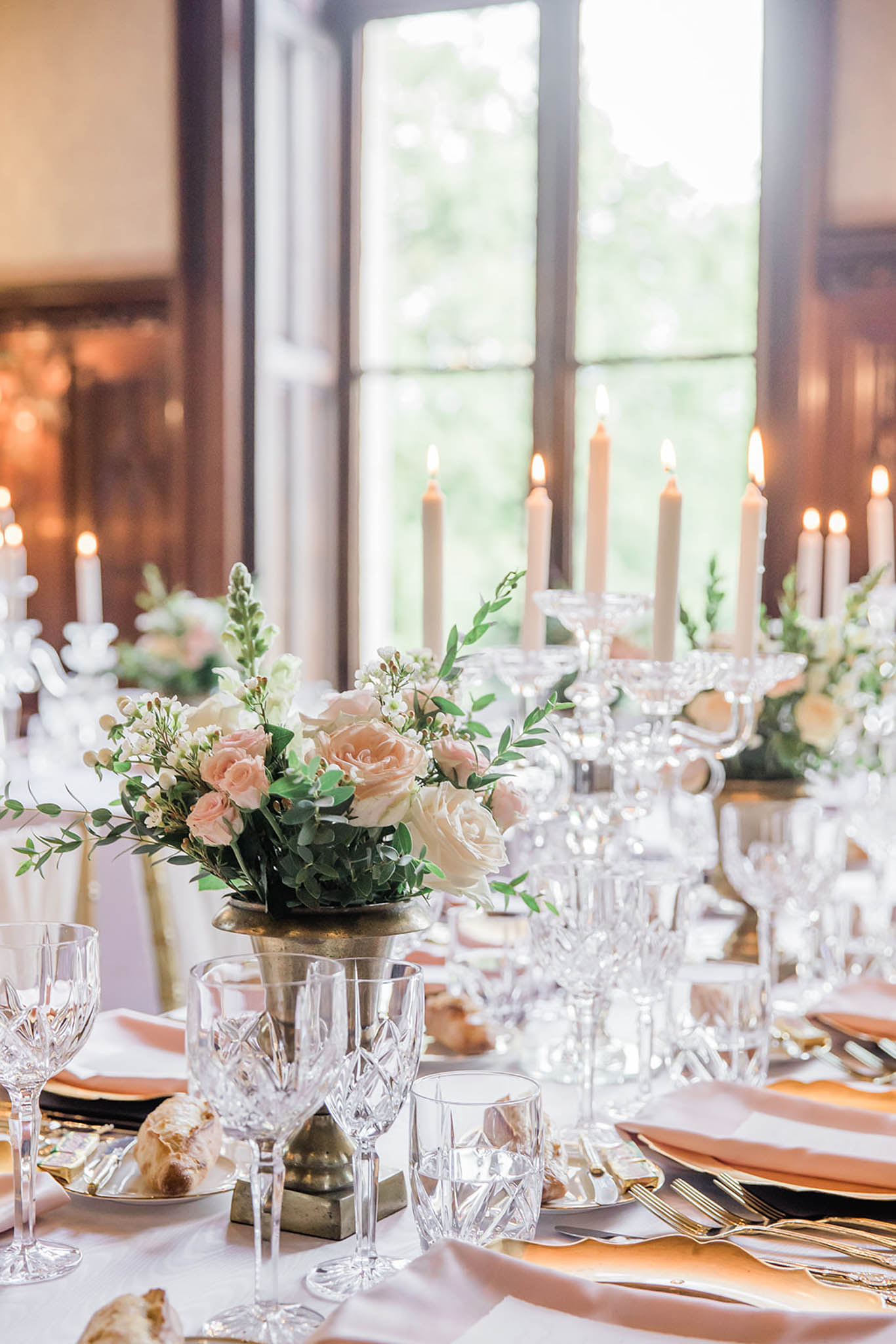 Reception table with blush roses in brass urn, crystal candelabras, gold flatware, and black charger plates