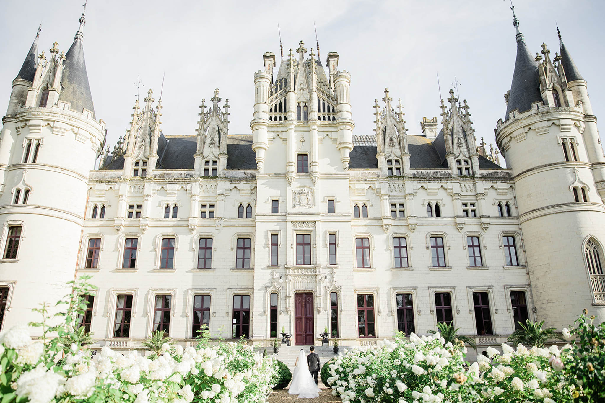 A wide-angle exterior shot of a large French Gothic-Renaissance château, featuring cream-white stone façade, dark slate-roofed pointed turrets, ornate carved stonework, and dark burgundy-painted doors and window frames. A bride in a long white gown with a cathedral-length veil and a groom in a dark suit stand together at the base of the château's grand entrance steps, viewed from behind, small in scale against the imposing three-story building. The foreground is filled with white flowering shrubs — likely white roses and white hydrangeas — creating a lush border that frames the couple. The composition is a centered wide shot taken from a slightly elevated angle, emphasizing the full architectural scale of the venue. Potential venue feature image.
