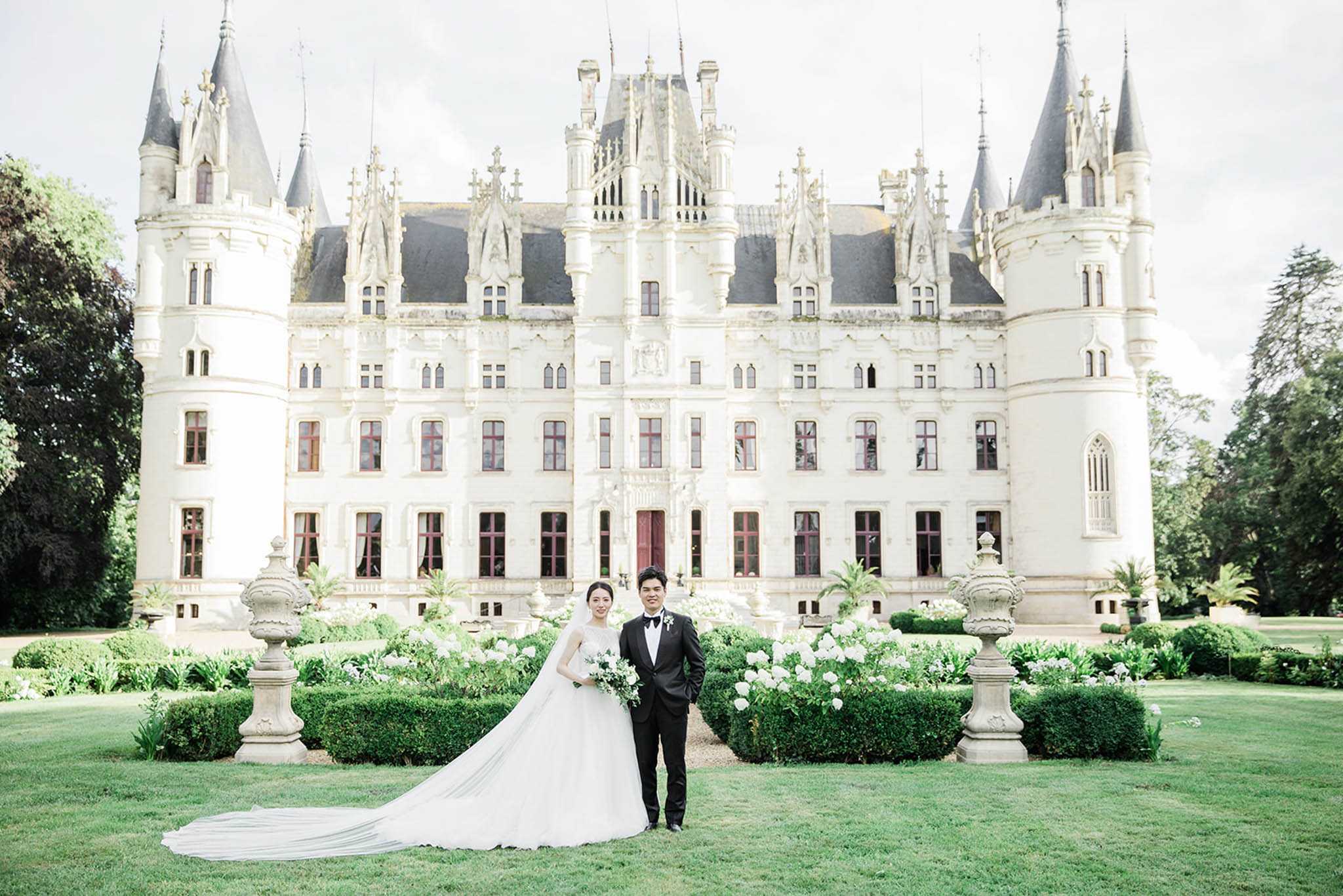 A couple portrait taken outdoors on the manicured lawn in front of a large white French Gothic-style château featuring pointed turrets, ornate stone detailing, and dark slate roofing. The bride wears a full-skirted ivory ball gown with a cathedral-length train and a long veil, holding a bouquet of white blooms and eucalyptus foliage; the groom wears a black tuxedo with a white dress shirt and bow tie. The formal garden between the couple and the château features clipped boxwood hedges, white flowering shrubs, and stone urn pedestals, creating a classic, symmetrical backdrop. This is a wide mid-distance portrait with the château centered behind the couple, shot in a clean, classic style with a white and green decor palette. Potential venue feature image.