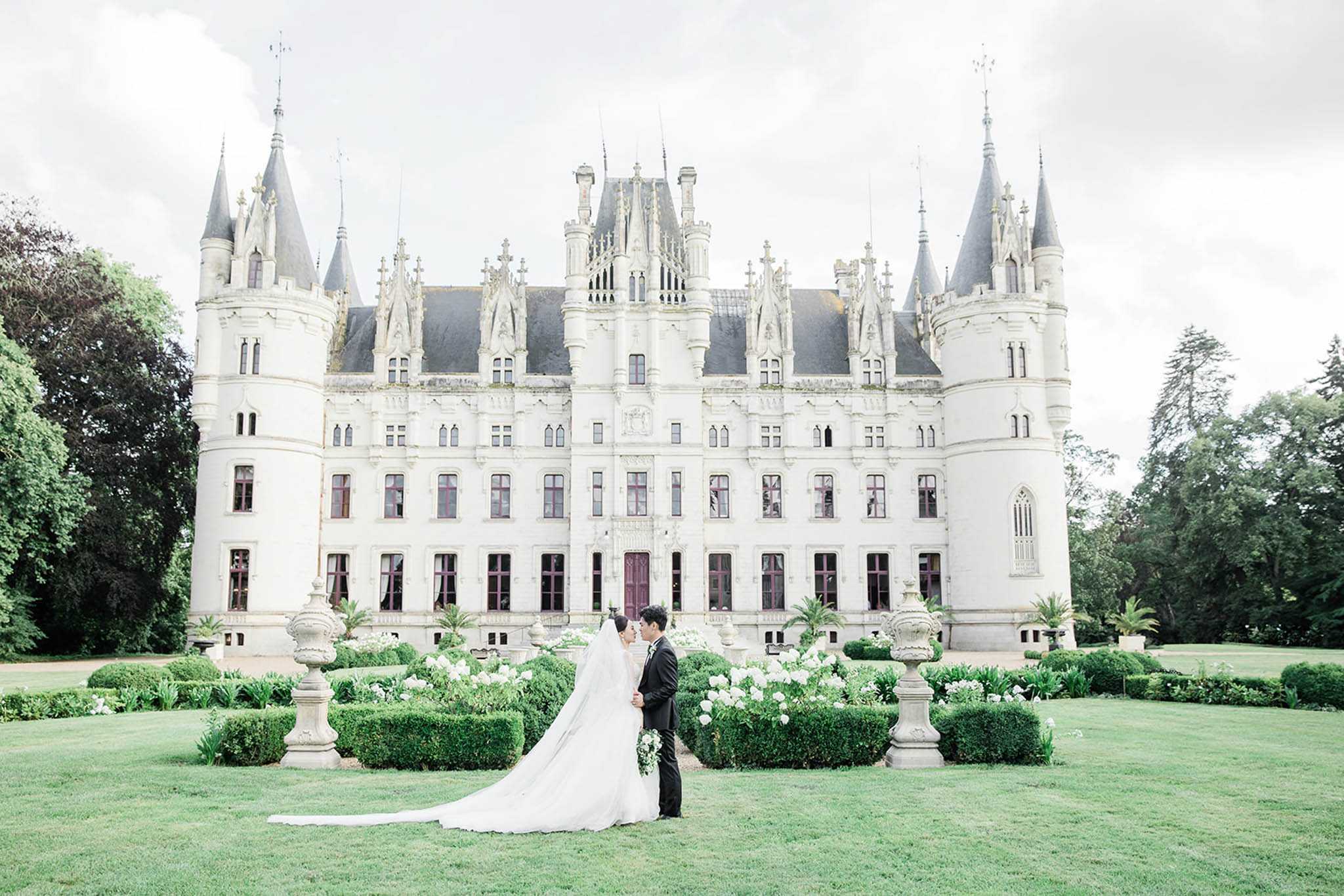 Bride and groom with foreheads touching on formal lawn before Gothic chateau with turrets and boxwood hedges