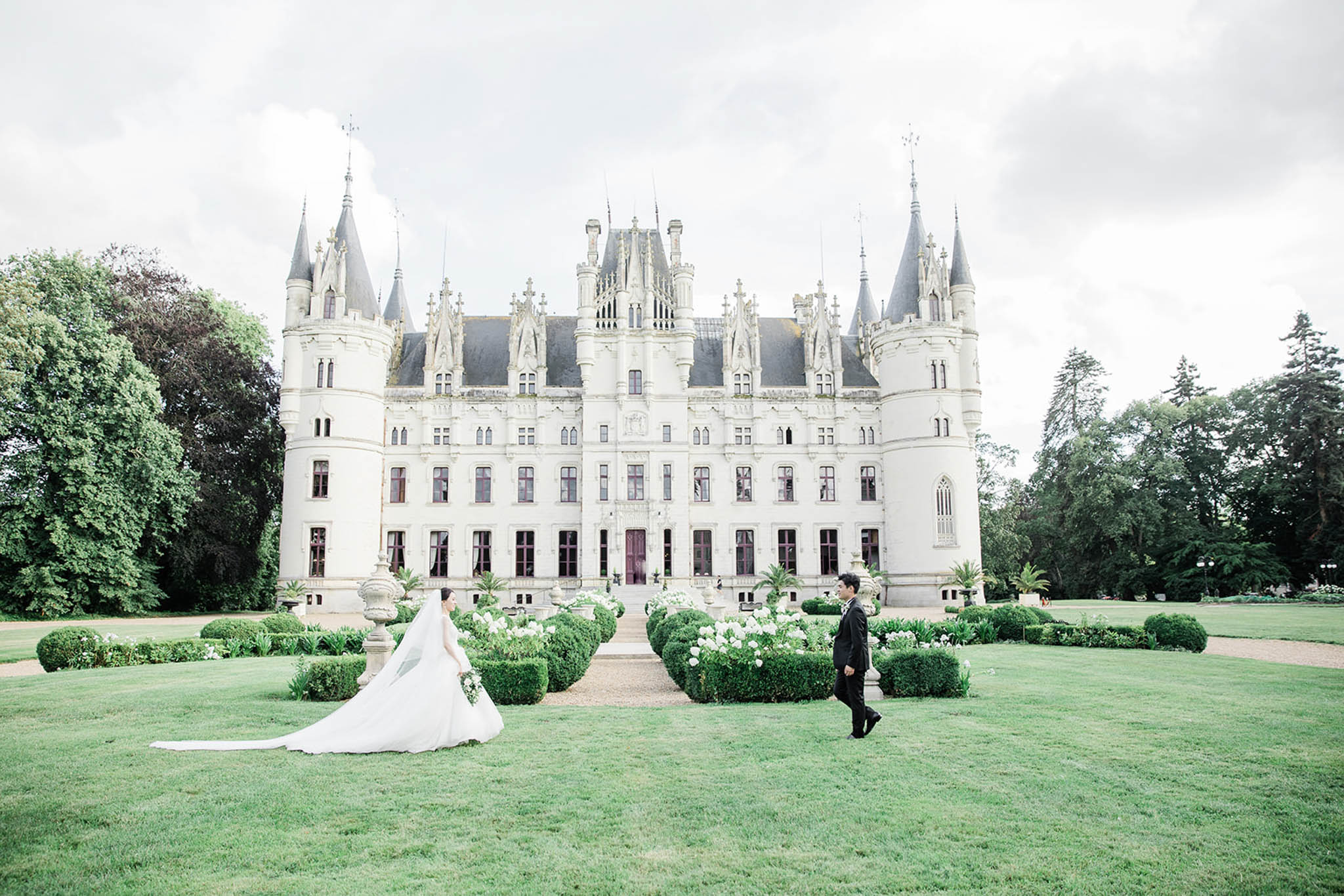 A couple portrait taken on the front lawn of a large French château featuring white limestone architecture with pointed turrets, Gothic-style ornamentation, and a formal entrance flanked by clipped boxwood hedges and white flowering plants. The bride wears a white ball gown with a very long cathedral veil that trails across the grass, and carries a small bouquet; the groom is dressed in a black tuxedo. The two are positioned apart on the lawn, facing each other at a distance, with the full château façade centered between them. The composition is a wide shot that deliberately places the couple small within the frame to emphasize the scale of the building and its formal gardens. Potential venue feature image.