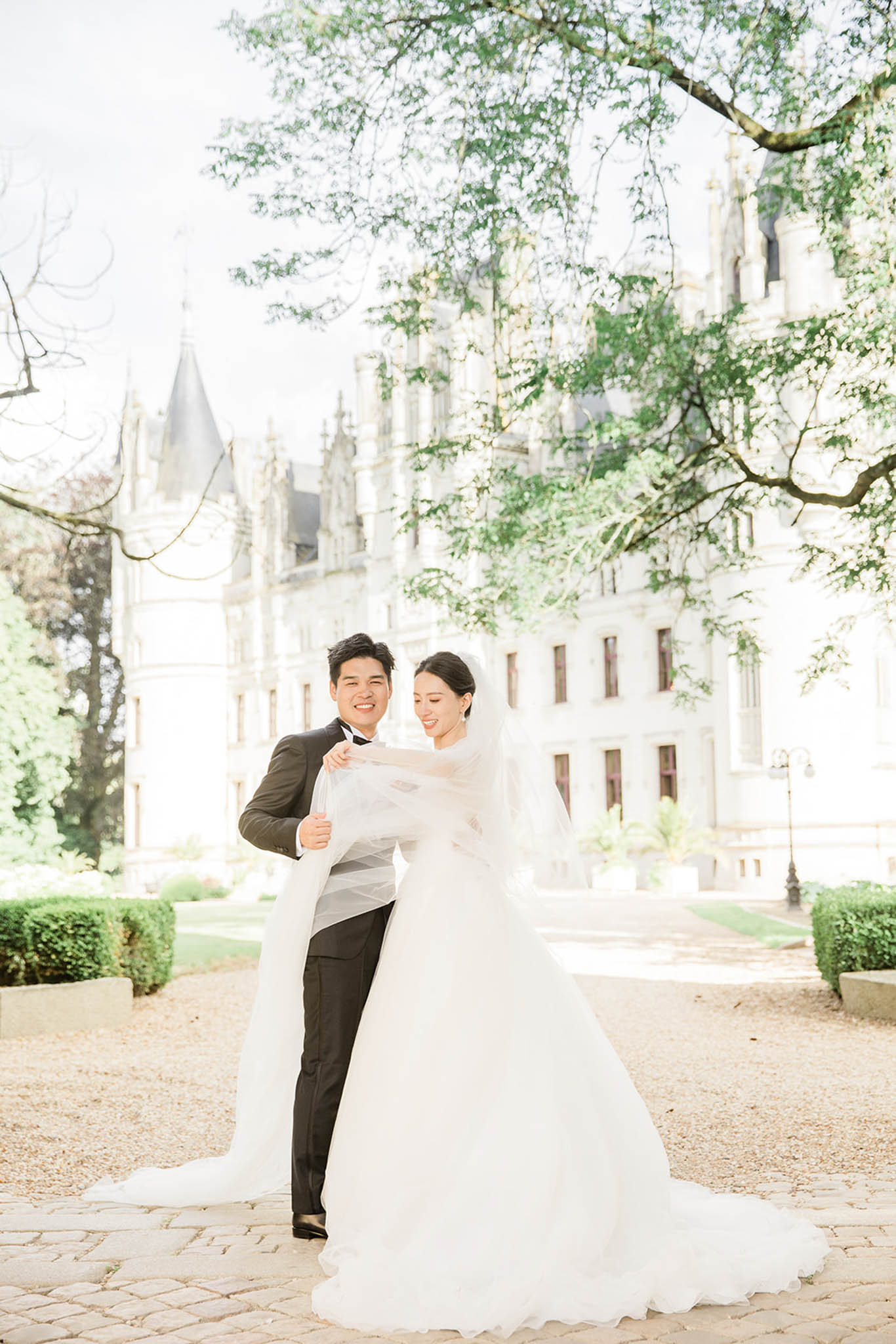 Groom lifting bride's cathedral veil in chateau grounds, both smiling, white ball gown and charcoal suit