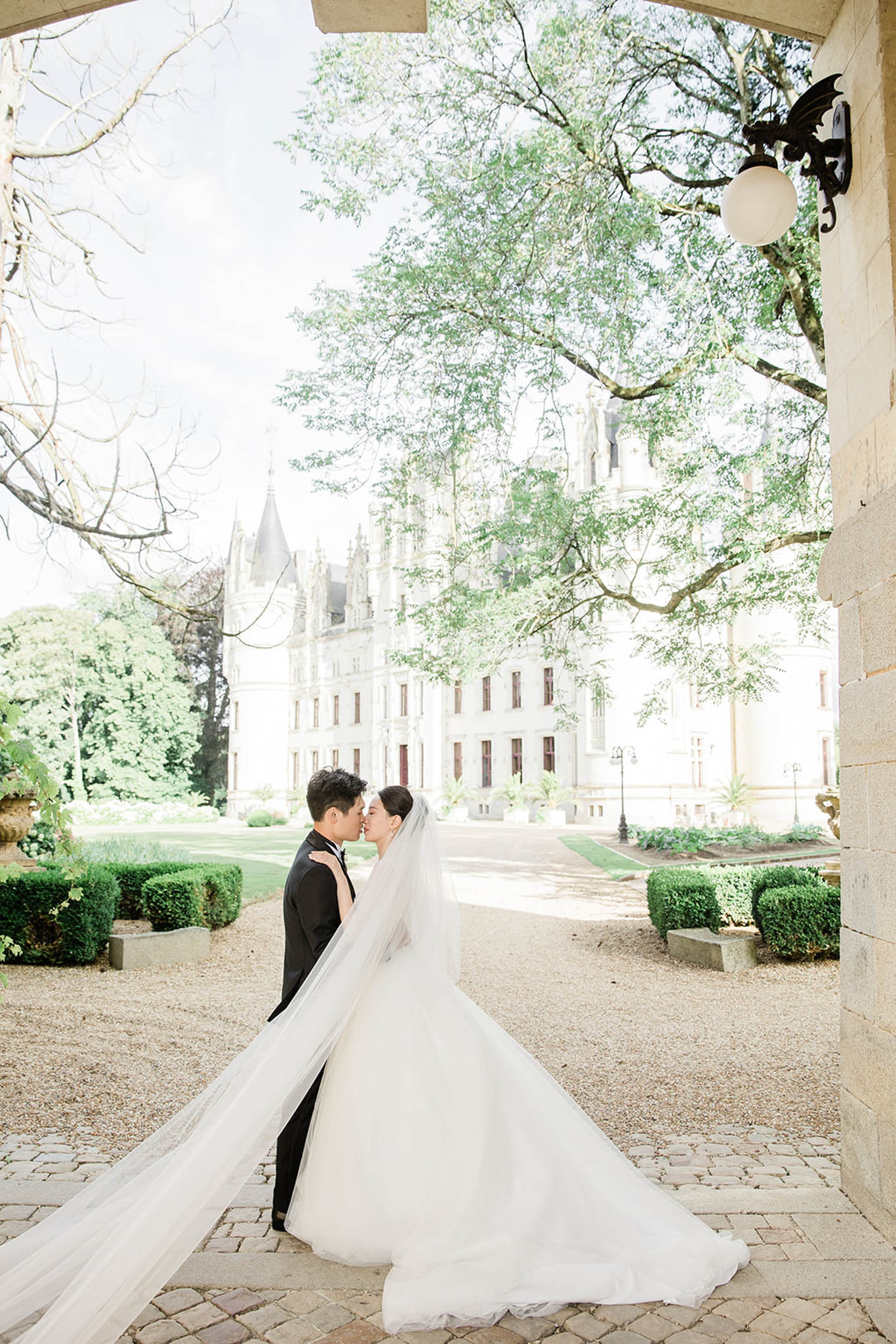 Bride in ballgown with cathedral veil and groom kissing in chateau courtyard framed by stone archway