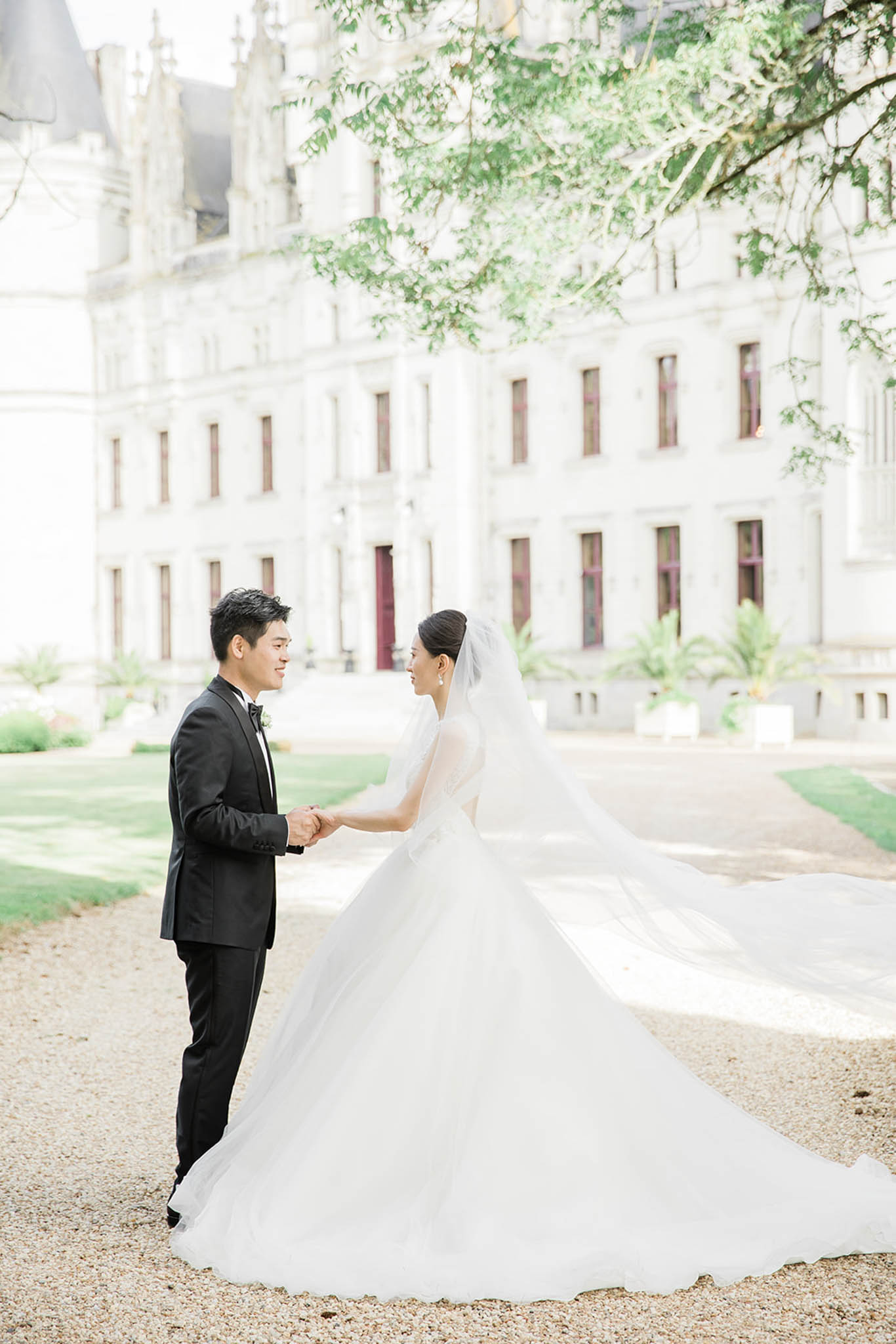 Bride in white ballgown with cathedral veil and groom in black tuxedo facing each other before chateau