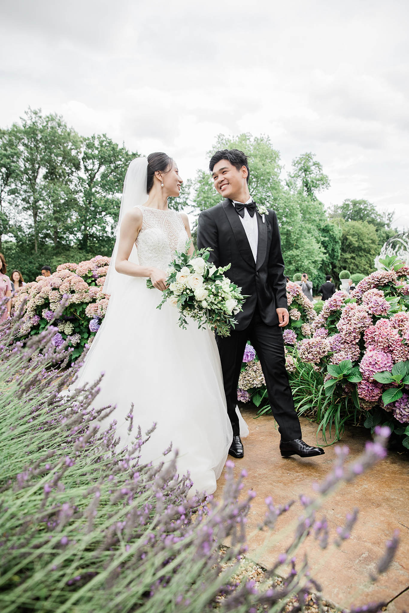 A couple walks together along a stone path in a formal garden, smiling at each other in a candid portrait-style shot. The bride wears a white A-line gown with a beaded sleeveless bodice and a long cathedral veil, carrying a loose bouquet of ivory garden roses, white ranunculus, and trailing greenery. The groom is dressed in a black tuxedo with a bow tie and a white boutonnière. The garden setting features large clusters of pink and mauve hydrangeas lining the path, with lavender plants in the foreground, and several guests are visible in soft focus in the background. The overall styling is classic and formal with a lush garden aesthetic.