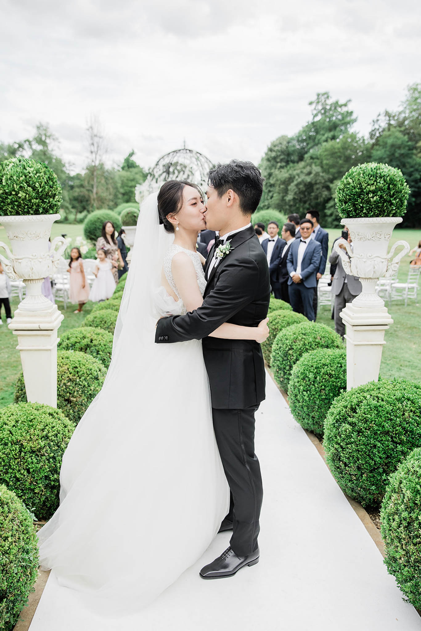 First kiss on white carpet aisle flanked by boxwood topiaries and stone urns in formal garden