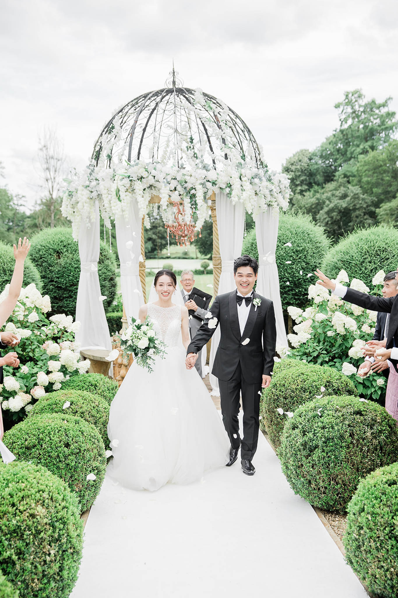 The bride and groom walk back down the aisle together after their outdoor ceremony, holding hands and smiling broadly while guests on either side throw white flower petals. The ceremony takes place in a formal French garden with manicured box hedges lining a white aisle runner and white hydrangea blooms flanking the path. Behind the couple stands an ornate wrought-iron dome gazebo draped with white fabric panels and adorned with cascading white roses and orchids, with a pink crystal chandelier hanging at its center. The bride wears a full ballgown with an illusion lace bodice and carries a bouquet of white roses and eucalyptus; the groom wears a black tuxedo with a black bow tie and a white floral boutonnière. The overall decor palette is white and green with a classic, formal style. Wide portrait shot taken from a low angle looking up the aisle.