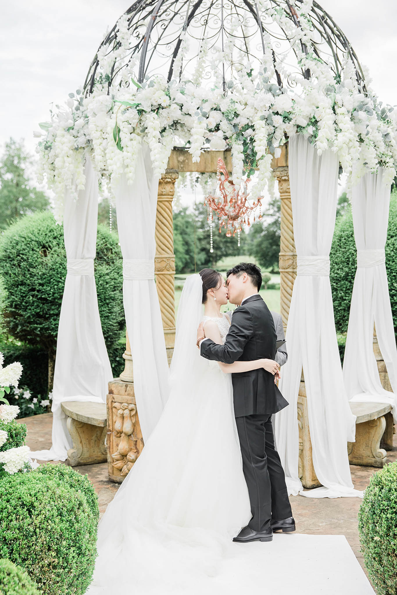 A couple shares a kiss during an outdoor ceremony beneath a stone and wrought-iron domed gazebo structure. The bride wears a white ballgown with a lace bodice and a long cathedral veil, while the groom wears a dark charcoal suit. The gazebo is decorated with cascading white florals — including white wisteria, roses, and eucalyptus — draped across the dome, with white fabric panels tied to the stone columns on each side. A rose-gold chandelier with crystal drops hangs at the center of the structure. Stone benches with carved figural bases flank the columns, and a white aisle runner leads toward the couple. The surrounding formal garden features neatly trimmed boxwood hedges and topiaries, with white hydrangeas visible at the edges of the frame. The overall decor palette is white and green with a classic, formal French garden aesthetic. Full-length portrait shot with the couple centered in the frame.