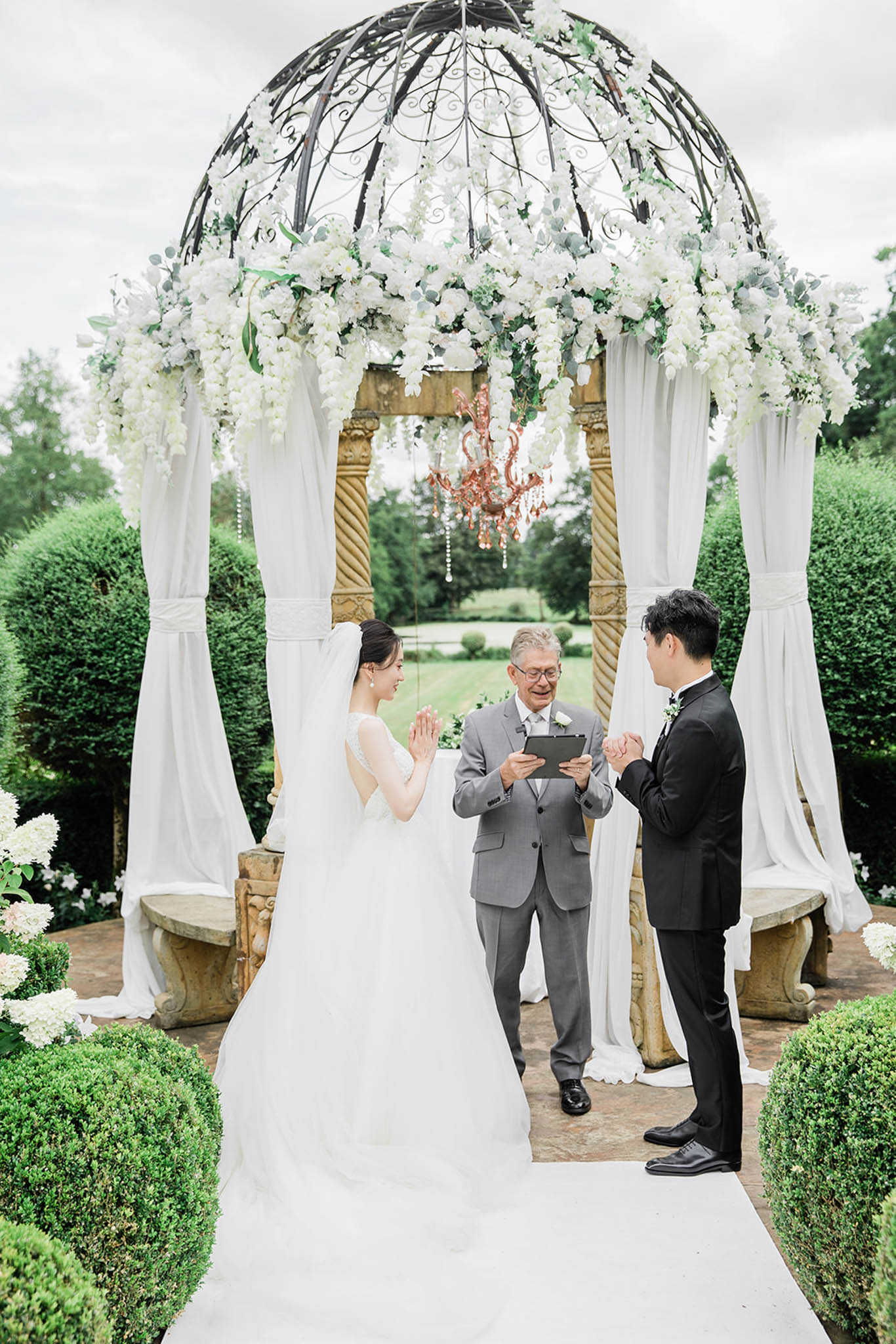 Couple holding hands under iron gazebo with white roses, crystal strands, and rose-gold chandelier