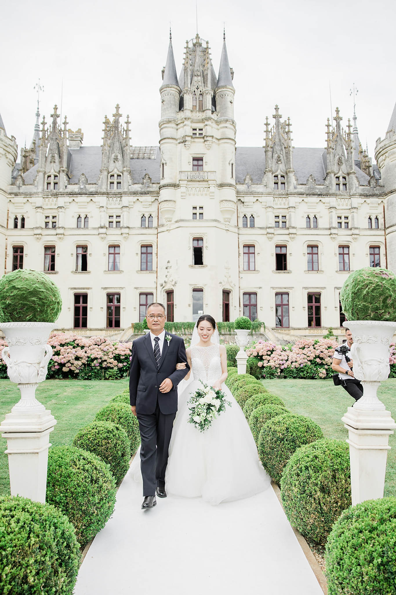 A bride is being walked down a white aisle runner by an older man, likely her father, during an outdoor ceremony processional at a French château. The bride wears a full ballgown with an embellished illusion neckline bodice and a long cathedral veil, carrying a cascading bouquet of white blooms and dark green foliage. The château facade features Gothic-Renaissance architecture with pointed turrets, ornate stone detailing, and red-trimmed windows, flanked by pink hydrangea borders and formally clipped box hedging with large white stone urns. A videographer is visible to the right of frame capturing the procession, and the composition is a wide portrait shot taken from ground level along the central garden axis. Potential venue feature image.