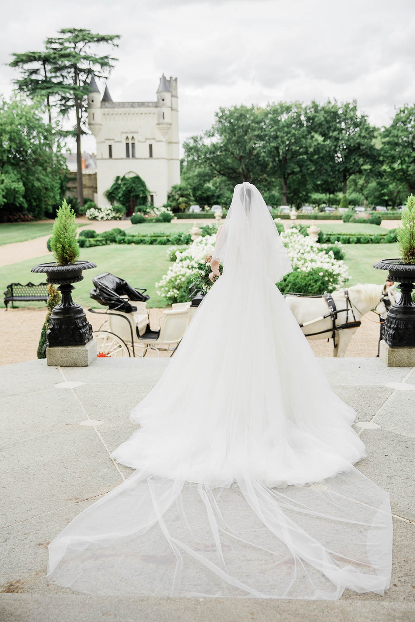 Bride from behind in ball gown with cathedral veil on terrace facing formal chateau garden with horse-drawn carriage