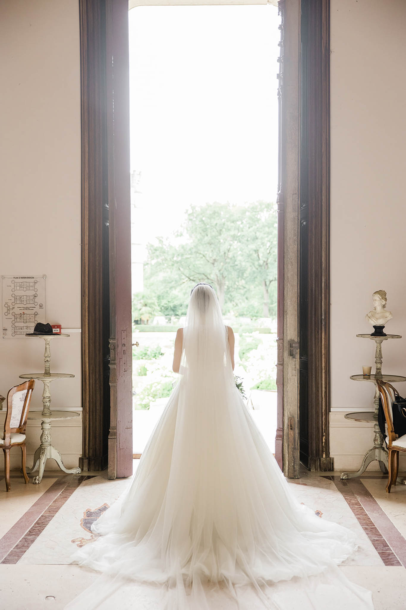A bride stands facing away from the camera in the entrance hall of what appears to be a French château, framed by tall, wide-open dark wood double doors that open onto a formal garden. She is wearing a full-volume ivory ballgown with a cathedral-length train that spreads across the marble-inlaid floor, paired with a long single-tier veil and a delicate headpiece. The interior space features cream-painted walls, ornate gilt-legged side tables, a classical marble bust on a pedestal to the right, a Louis XV-style chair, and a fire safety plan posted on the left wall. The composition is a full-length portrait shot from behind, centered symmetrically in the doorway, with the bright garden providing strong backlight that highlights the layers of the tulle gown and veil.