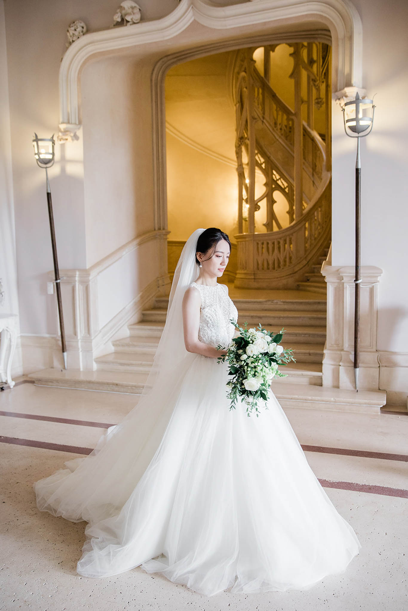 A bridal portrait taken indoors at what appears to be a French château or hôtel particulier, featuring an ornate carved stone grand staircase with wrought-iron balustrade in the background, warmly lit by wall-mounted lantern sconces. The bride stands alone in the foreground, wearing a full ballgown with a lace-embroidered bodice, voluminous ivory tulle skirt with a long train, and a cathedral-length veil. She holds a loose, garden-style bouquet of white roses, white filler florals, and abundant green eucalyptus and foliage. The marble floor features inlaid terracotta-toned geometric detailing. The composition is a three-quarter-length portrait shot with the staircase centered behind her as a key architectural backdrop. Potential venue feature image.