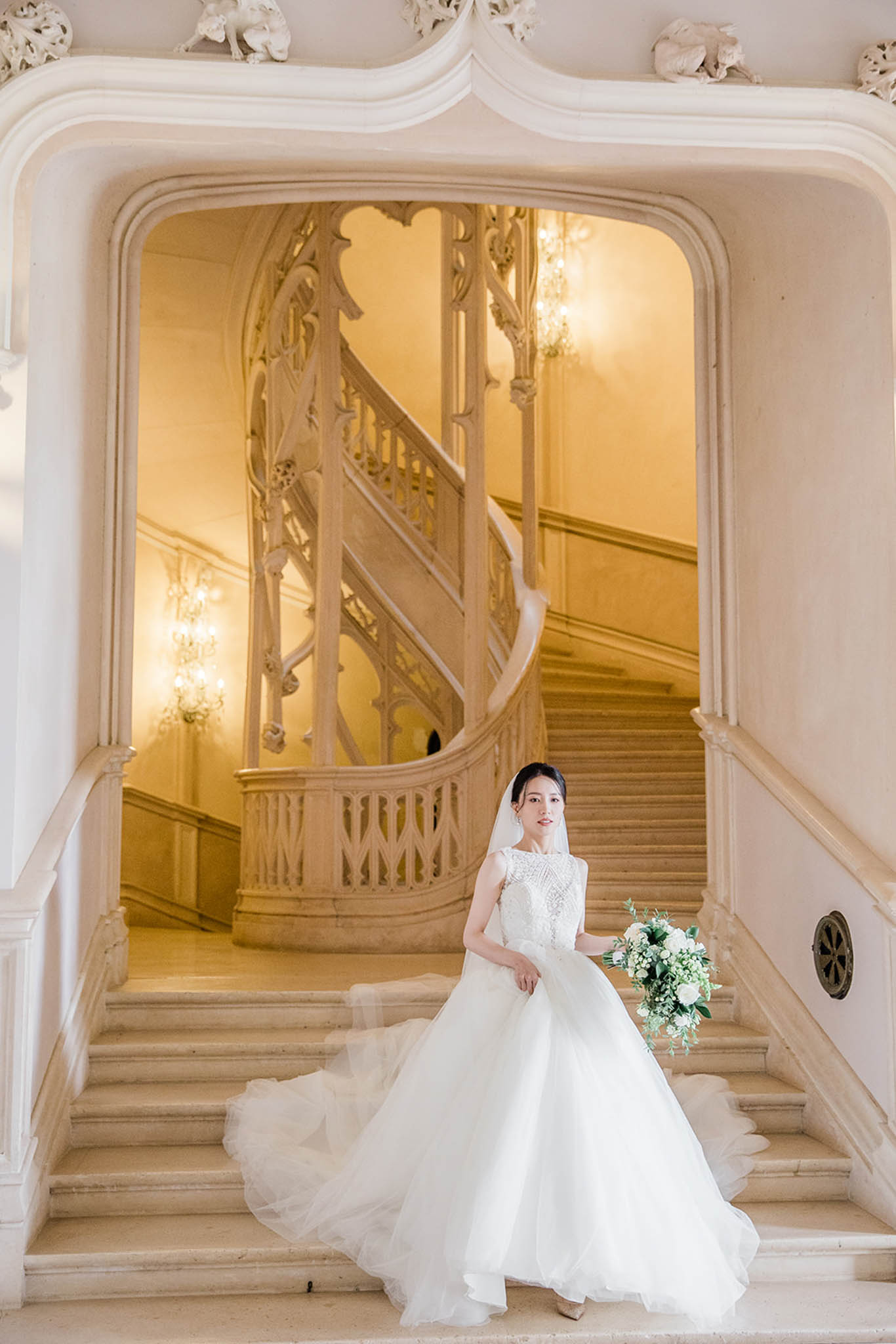 Bride in white ball gown with beaded bodice and cathedral veil on marble staircase inside ornate chateau interior