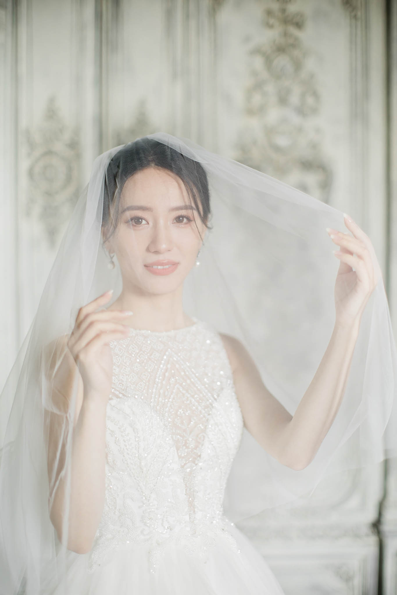 Bride in beaded ball gown lifting sheer tulle veil against white panelled wall in French chateau interior