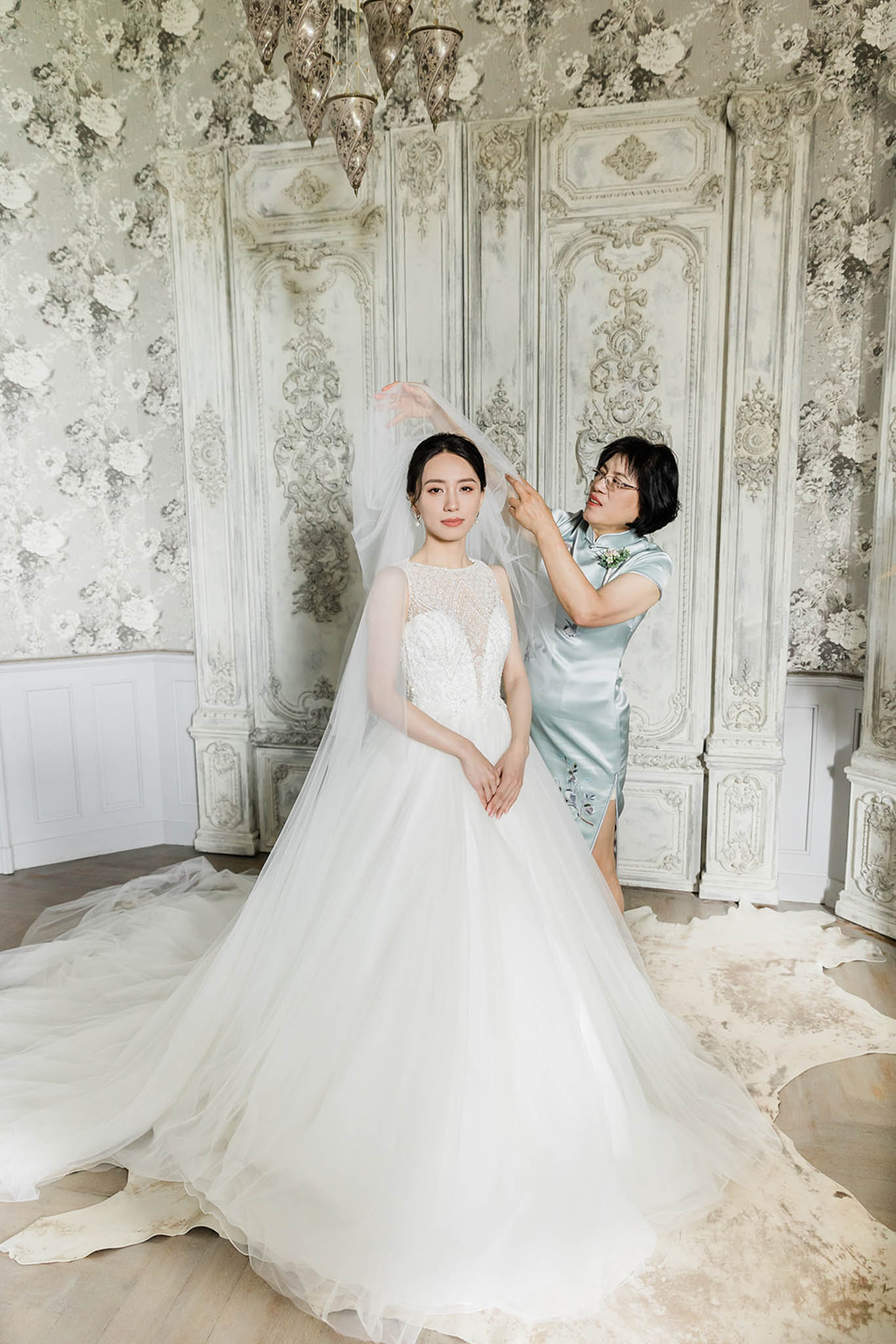 A getting-ready moment indoors, showing a bride standing while an older woman — likely her mother — adjusts her cathedral-length veil. The bride wears a full ballgown with a beaded illusion bodice and a voluminous white tulle skirt with an extended train; her hair is pulled back in a sleek updo with pearl drop earrings. The older woman is dressed in a pale blue satin qipao (cheongsam) with floral embroidery detail and a small corsage. The room features ornate white rococo-style carved panel screens, grey-and-white floral wallpaper, a Moroccan-style metal chandelier overhead, and a cowhide rug on light wood flooring, creating a classic French interior setting. The composition is a full-length portrait shot with both figures centered against the decorative backdrop.