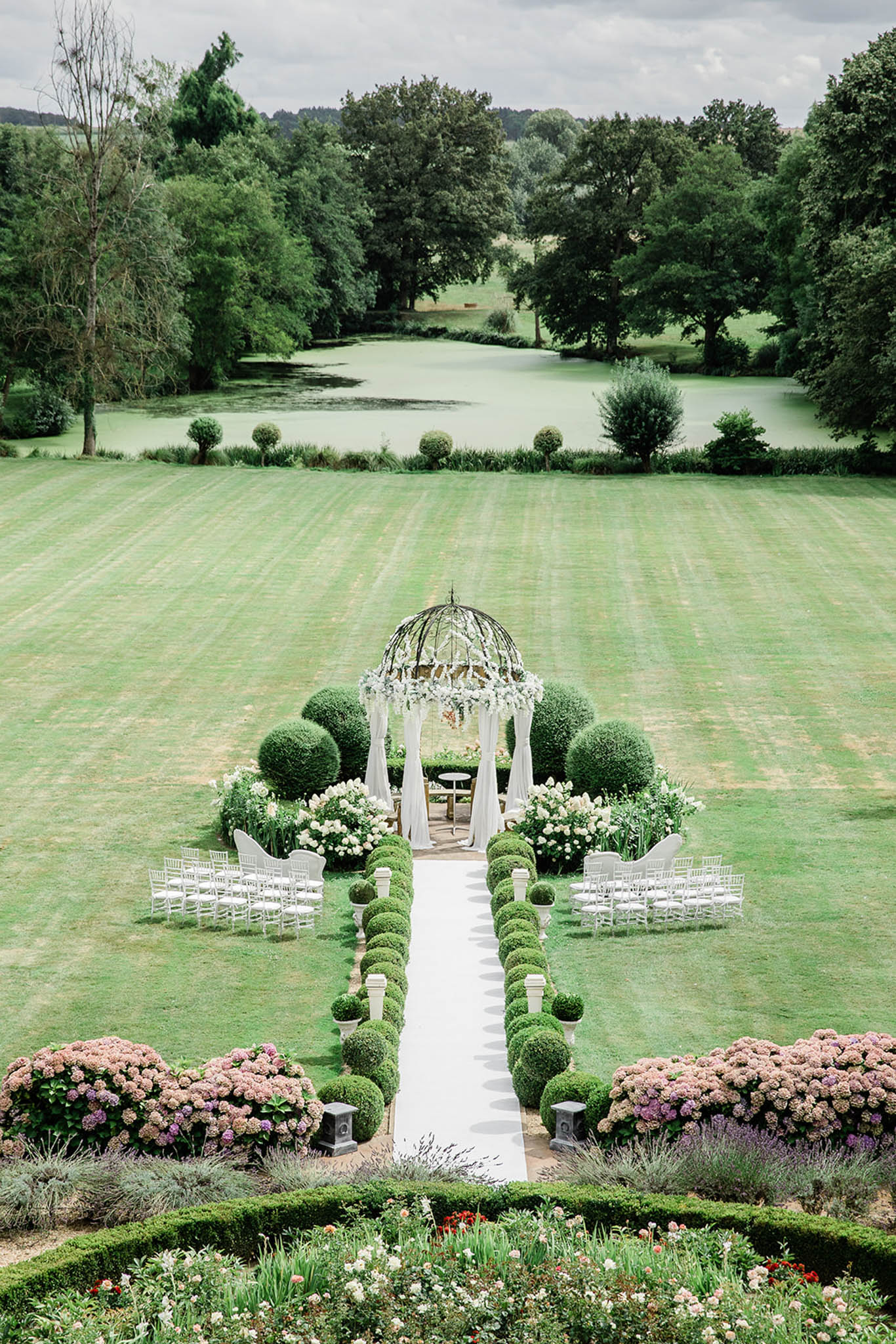 An aerial wide shot of an outdoor wedding ceremony setup in a formal French garden, with no guests or couple present. A white aisle runner leads toward a wrought-iron domed gazebo draped with white fabric curtains and topped with white floral arrangements, flanked by clipped spherical boxwood topiaries and large arrangements of white hydrangeas and garden roses. White chiavari chairs are arranged in rows on either side of the aisle, accommodating approximately 60–80 guests. In the foreground, formal parterre hedging borders beds of pink and mauve hydrangeas, lavender, and mixed blooms in cream and red. The grounds extend to a large ornamental lake bordered by clipped lollipop-standard trees. The overall styling is classic French formal garden with an all-white and soft blush floral palette. Potential venue feature image.