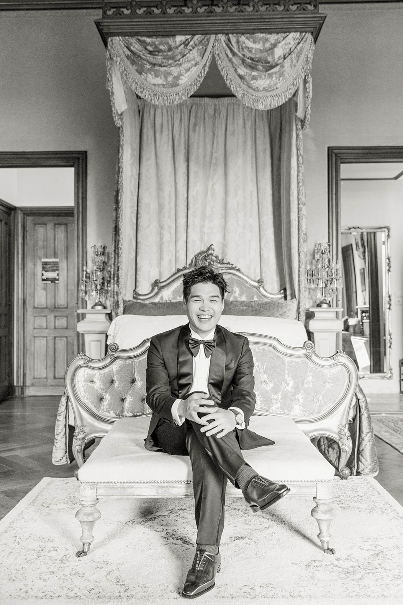 Black and white portrait of groom laughing in tuxedo seated on ornate canopied bed in baroque chateau room