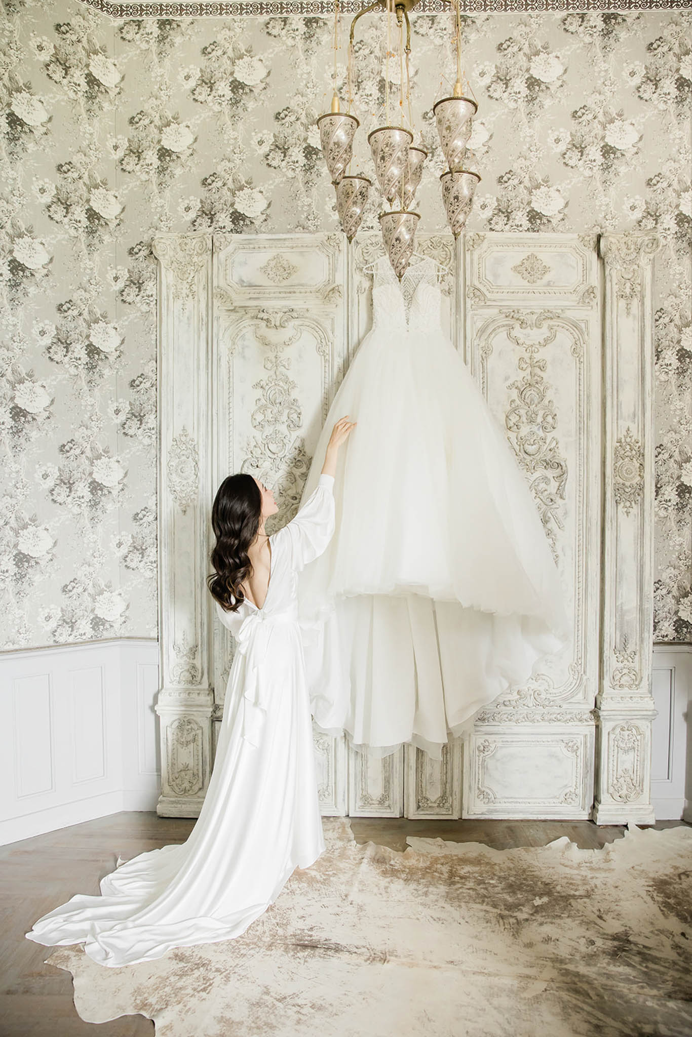A getting-ready scene shot indoors, showing a bride in a white silk long-sleeve bridal robe with a train reaching up to touch her wedding dress, which is hanging from a gold ornate chandelier. The wedding gown is a full ballgown silhouette in ivory with lace detailing at the bodice and a voluminous tulle skirt. The bride has long dark wavy hair and is viewed from behind. The room features grey and white large-scale floral wallpaper, a distressed white carved wood decorative panel behind the dress, and a cowhide rug on the hardwood floor, giving the space a French romantic interior styling. The composition is a full-length portrait shot that emphasizes both the robe and the hanging gown against the ornate backdrop.