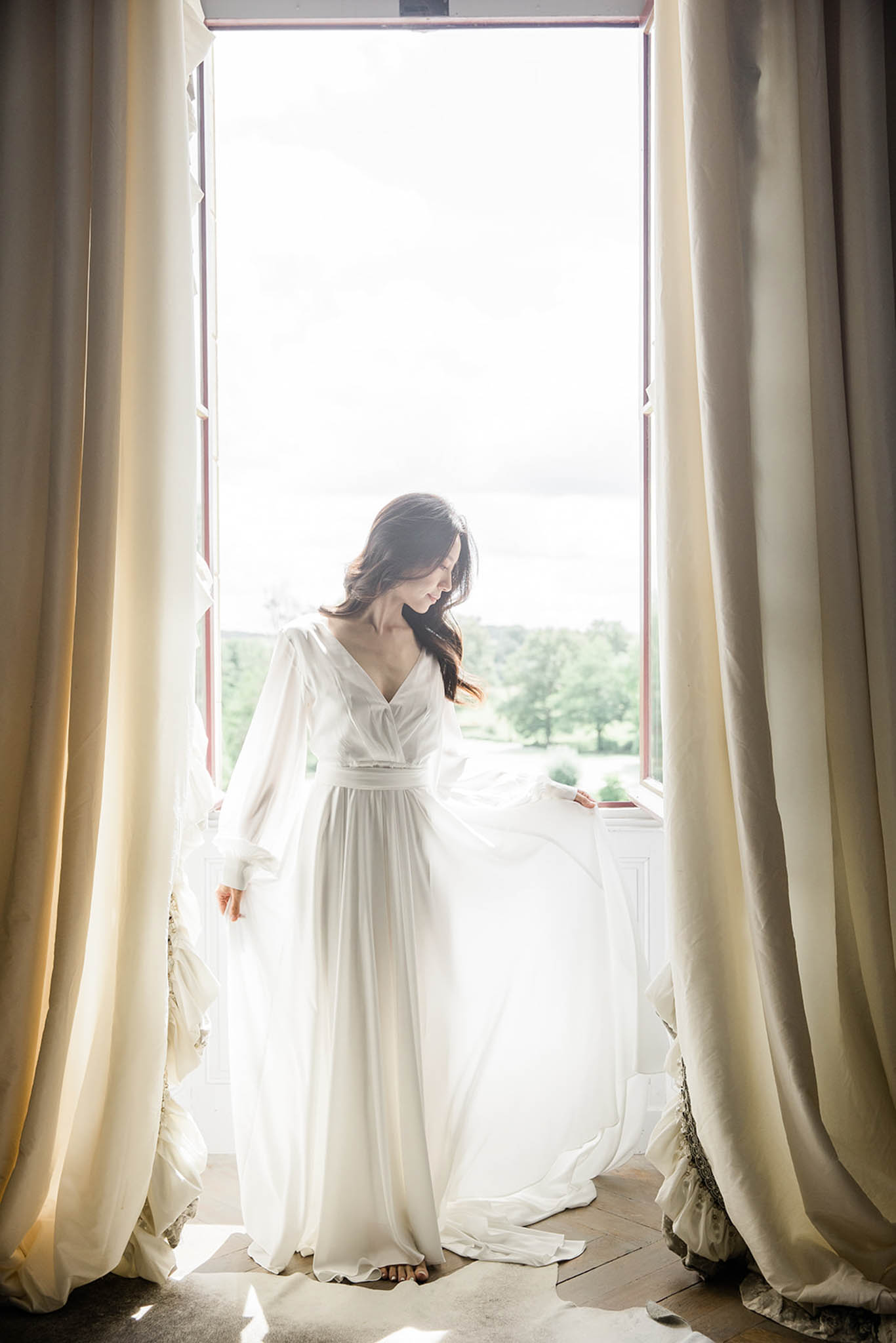 A bride stands in front of a tall open window in an indoor room, backlit by natural light flooding in from outside. She is wearing a floor-length white chiffon gown with long billowing sleeves, a V-neckline, and a gathered waist, and she looks downward as the skirt moves slightly in the breeze. The window is framed by floor-length cream curtains, and the wooden floorboards are visible beneath her feet. The composition is a full-length portrait shot, with the bride centered between the curtains and the overexposed outdoor landscape visible through the window behind her.