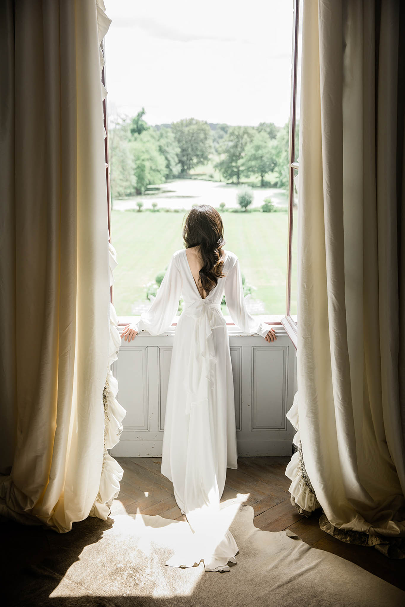 A bride stands with her back to the camera, looking out through tall open French windows toward a manicured parkland landscape. She is wearing a white long-sleeve wedding dress with a deep V-back, bow detail at the waist, and a flowing train that pools on the herringbone parquet floor. Her dark brown hair is styled in a loose side braid. The room features floor-to-ceiling cream linen curtains framing the window, and the interior walls are painted pale grey with wood panelling. Natural light streams in strongly through the window, casting a bright patch across the train on the floor. The shot is a full-length portrait taken from behind, emphasizing the back detail of the dress and the contrast between the interior and the green grounds outside.
