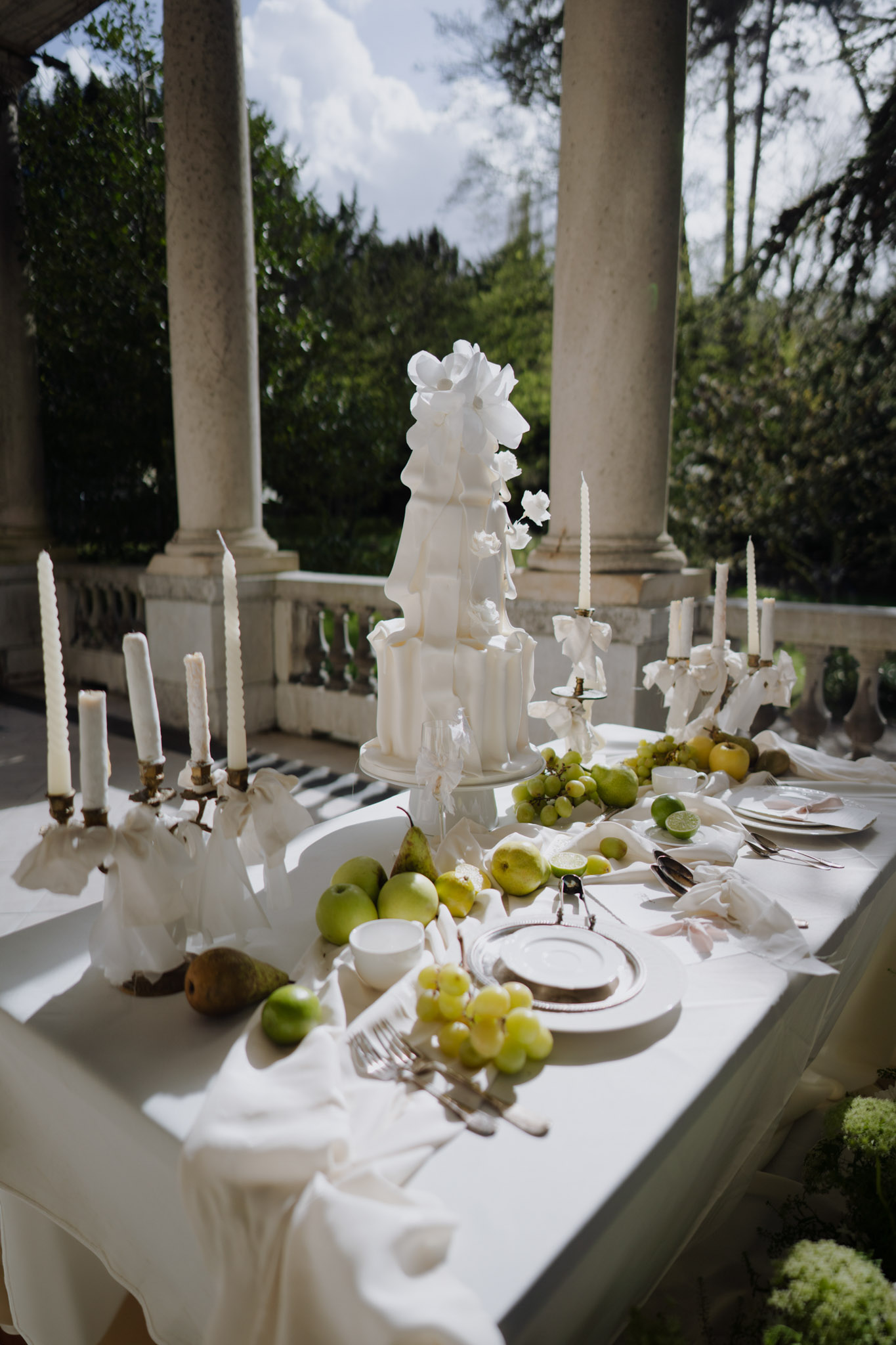 Three-tier ruffled white cake on fruit-scattered table with brass candelabras on columned terrace