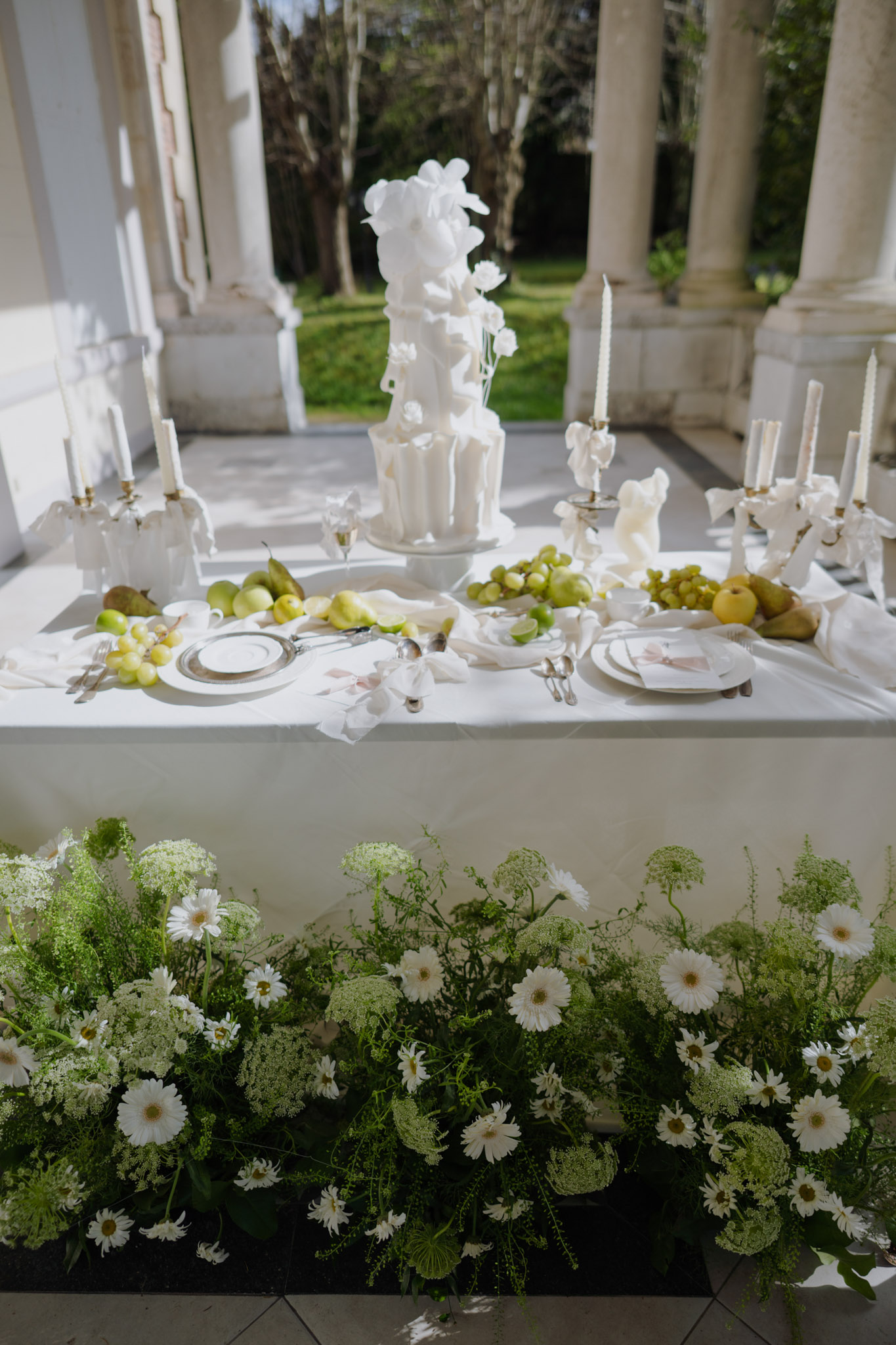 All-white tiered cake with sugar orchids on colonnade terrace beside green fruit and gerbera ground display
